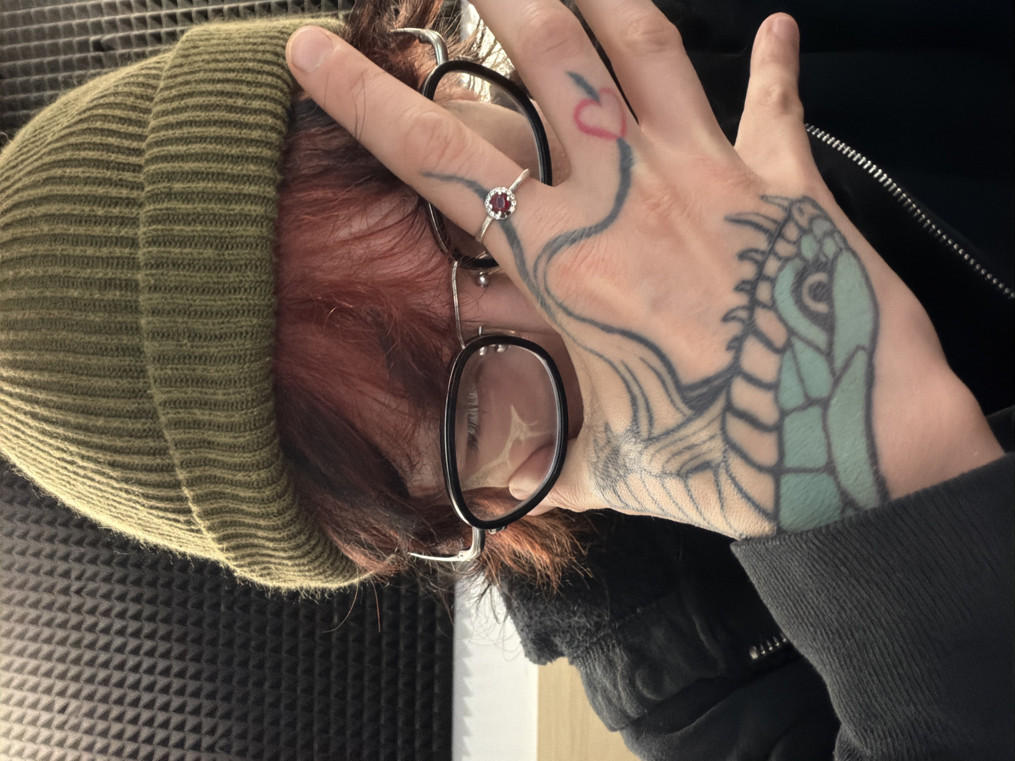 A person is depicted indoors against a soundproofing-textured wall. The individual wears a ribbed, olive-green beanie, black-rimmed glasses, and a dark hooded jacket. They have red hair and fair skin. The left hand is raised, partially obscuring the face and holding the glasses' frame. Visible tattoos are present on the back of the hand and fingers, including a heart shape and a stylized aquatic creature. A silver ring with a small red stone adorns a finger. The scene's exact location within Sofia, Bulgaria, cannot be determined from the image alone.