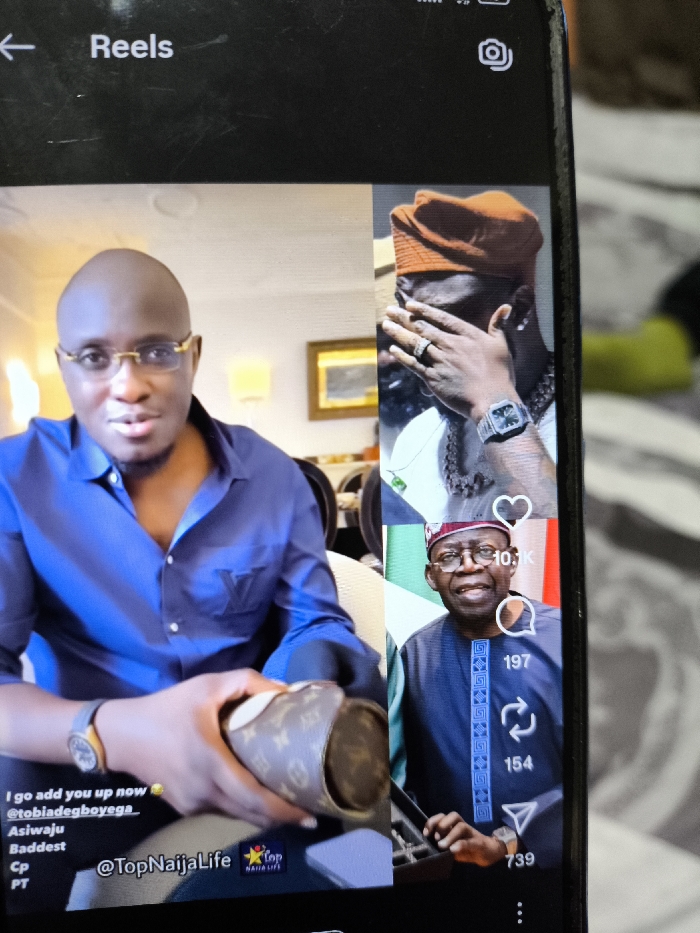 A mobile phone screen, displaying a social media application's "Reels" interface, is captured in a slightly blurred photograph. The phone's top left shows a back arrow and the word "Reels," while the top right has a camera icon and network/battery indicators. The screen is vertically split, presenting three distinct video or image frames.
On the left, a bald, light-skinned man with gold-rimmed glasses and a blue collared shirt, featuring a subtle "LV" (Louis Vuitton) logo on the left chest, looks directly forward. He wears a dark-strapped watch with an orange face on his left wrist. His hands are in the lower foreground, holding a brown cylindrical object patterned with the Louis Vuitton monogram. Text overlays include "I go add you up now," "@tobiadegboyega," "Asiwaju," "Baddest," "Cp," "PT," and "@TopNaijaLife."
The upper right frame shows a dark-skinned individual wearing an orange/brown head covering and a white garment. Their left hand is raised, covering part of their face, revealing a prominent silver ring on their ring finger. A silver-toned square-faced watch is on their left wrist, and a thick, gold-colored chain is visible around their neck.
The lower right frame depicts a smiling man with spectacles, wearing a blue traditional Nigerian attire (agbada or similar) with intricate patterns. He is positioned in front of a background featuring what appears to be the Nigerian flag. Social media engagement metrics are displayed: a heart icon with "10.1K," a comment bubble with "197," a share icon with "154," and a save icon with "739."
The phone itself exhibits minor scratches on its screen. The background surface upon which the phone rests or is held appears to be a patterned textile, possibly a duvet or blanket, in muted tones. The image provides no visual indicators of its physical location in Nicosia, Northern Cyprus / Cyprus.