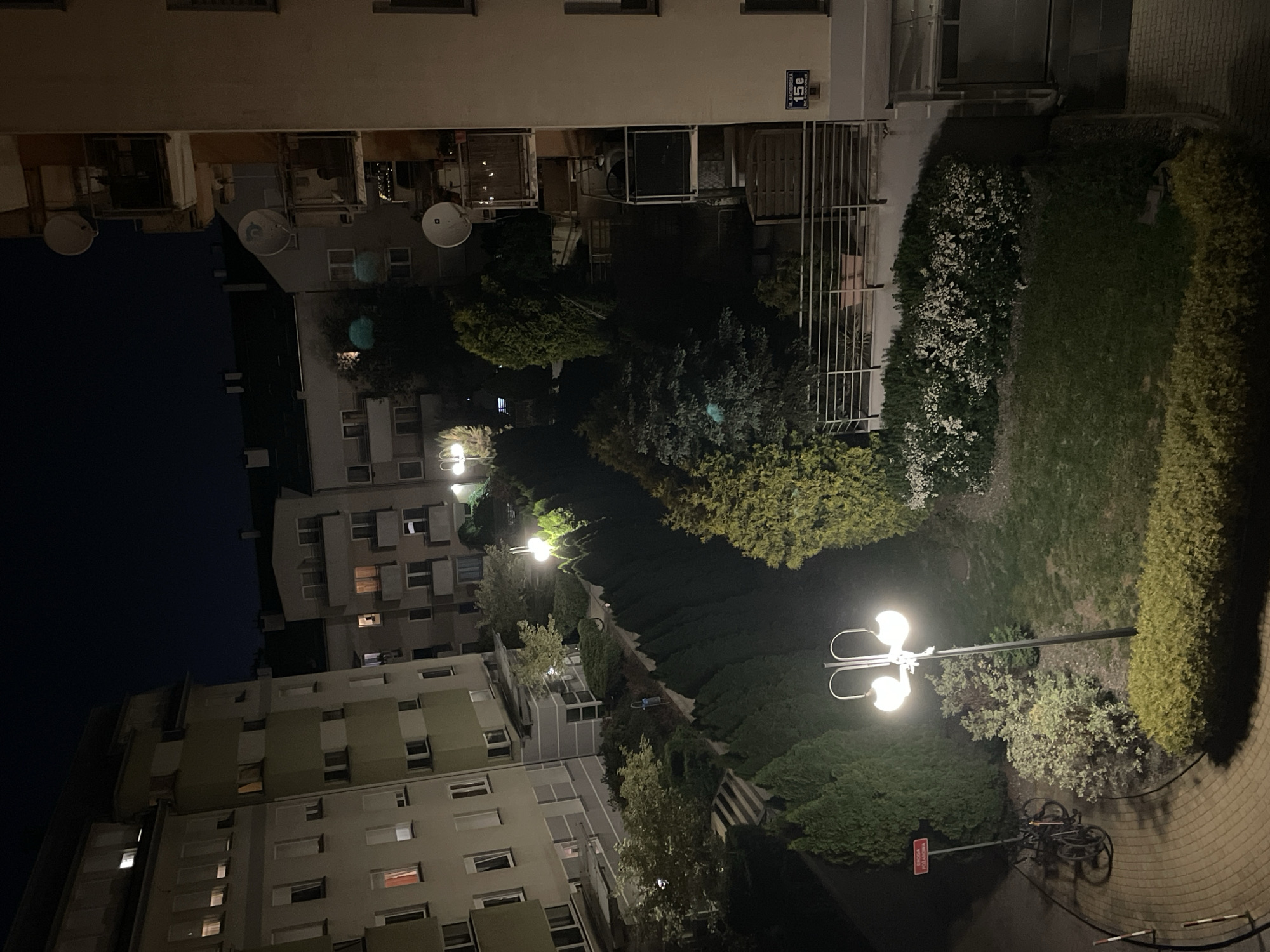 The image shows an outdoor scene at night. There are several buildings present, appearing as residential apartments, with windows indicating signs of occupancy. Streetlights illuminate a pathway lined with hedges and trees. A bicycle is parked near one of the lampposts. A sign with Polish text "Droga pożarowa" (Fire Road) is also visible. The location is Warsaw, Poland based on the address sign "Ul. Radziwie 15e".