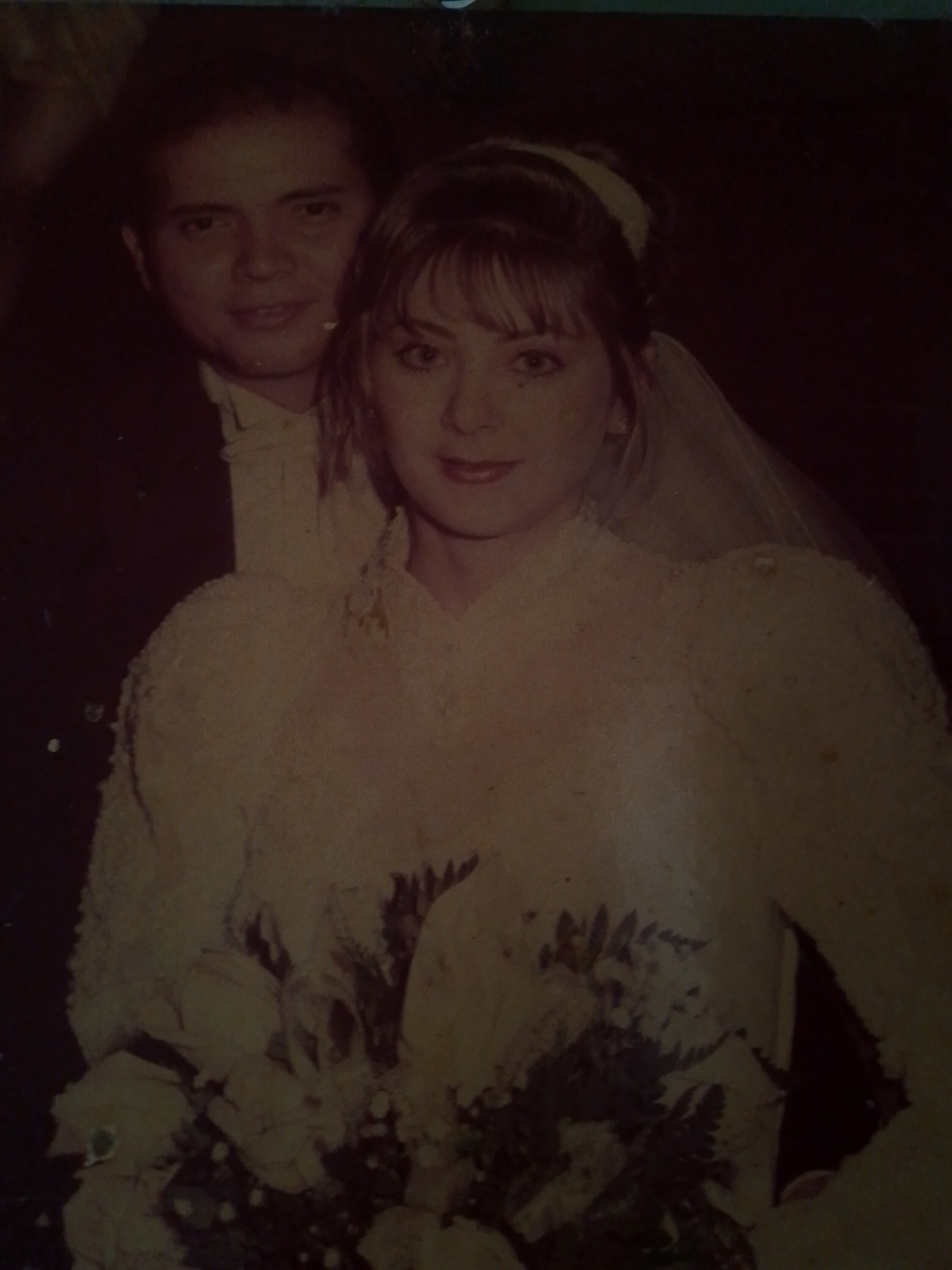 The image depicts a wedding portrait of a couple. The woman, presumably the bride, is wearing a white wedding dress with large, puffy sleeves and a veil. She has bangs and her hair is styled up. She is holding a bouquet of white flowers. Behind her is a man, presumably the groom, wearing a dark suit with a white shirt and bow tie. The background is dark and indistinct. It is impossible to determine if this picture was taken in Caracas, Venezuela.