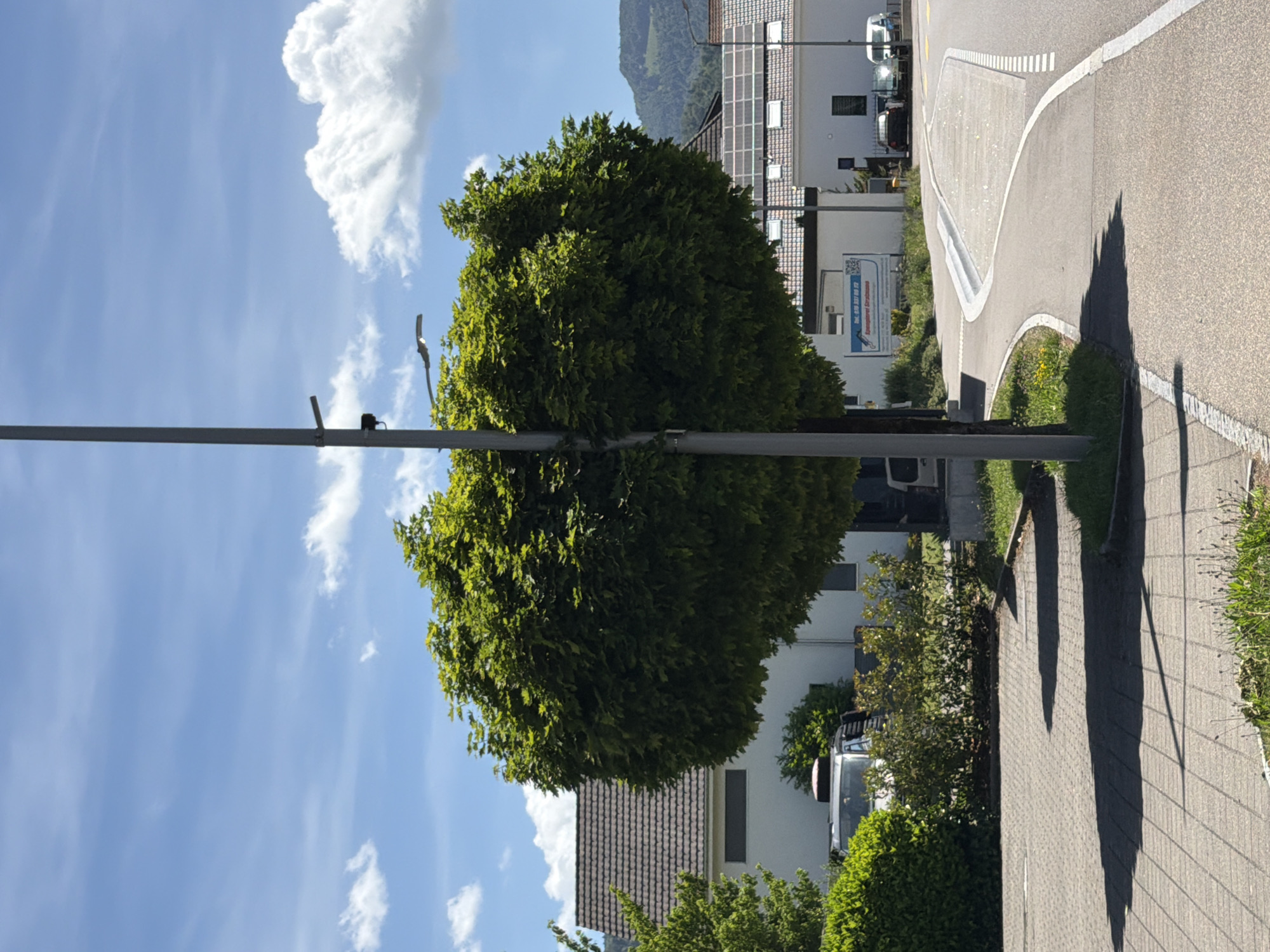 The image depicts a street scene on a sunny day with a bright blue sky and scattered white clouds. A large, round, leafy green tree dominates the foreground, casting a shadow on the paved ground. A grey lamppost stands in front of the tree, bisecting the frame vertically. In the background, several white buildings with dark tiled roofs are visible. Some of the roofs are equipped with solar panels. A paved road curves through the scene, bordered by sidewalks. A few parked cars are visible in front of one of the buildings. The overall impression is of a clean, modern urban or suburban environment. The location is identified as Unknown, Unknown.