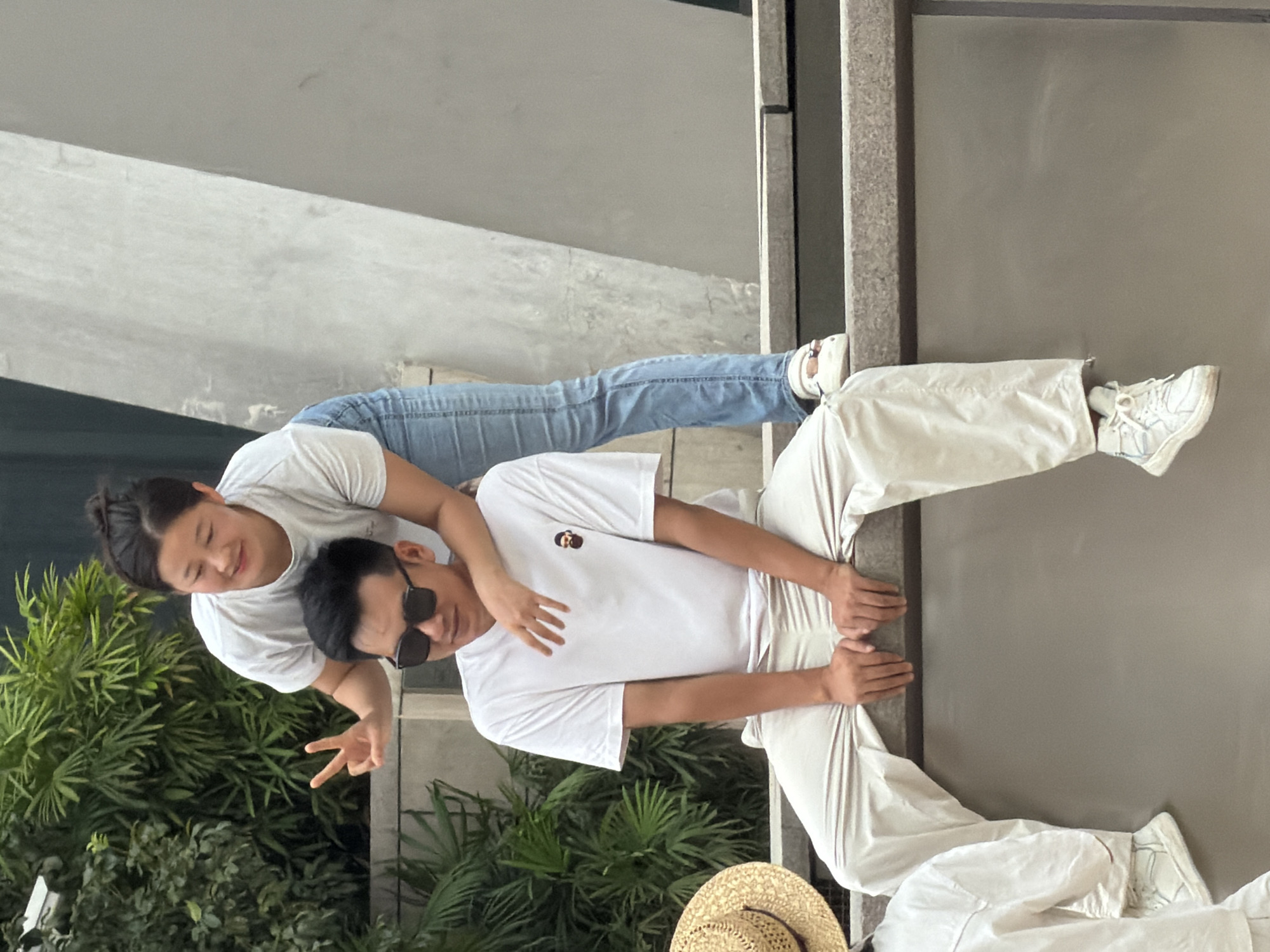 In Yuzhong District, China, two individuals are captured in a candid moment. A woman, standing behind a man, leans forward with her left arm around his neck and her right hand touching his shoulder, her fingers splayed. She is wearing a white t-shirt and blue jeans, her hair pulled back in a bun, and she is smiling. The man, seated on a ledge, wears a white t-shirt with a small brown graphic and loose-fitting beige pants. He has dark hair, is wearing sunglasses, and is looking downwards. His hands rest on his knees. The environment appears to be outdoors, with lush green foliage visible behind them and a textured, concrete structure to their right. A straw hat sits on the ledge to the left of the man.
