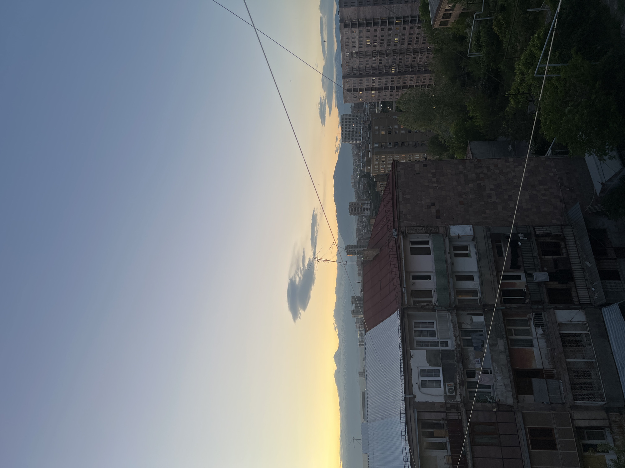 This is a high-angle view taken in Yerevan, Armenia, likely from the rooftop of a building. The image is oriented vertically with the sky occupying the left and upper portions of the frame. The sky transitions from a blue hue to a yellow-orange gradient near the horizon, indicative of either sunrise or sunset.
Buildings dominate the lower and right portions of the image. Several aging apartment buildings with a mix of exposed brick, whitewashed walls, and visible decay are present. Some apartments have windows missing or partially covered, while others have air conditioning units installed.
In the background, there are taller, Soviet-era apartment blocks stretching into the distance. The horizon is marked by mountains. Thin wires, likely power or communication lines, crisscross the upper portion of the frame. A few small clouds are visible in the sky near the horizon.