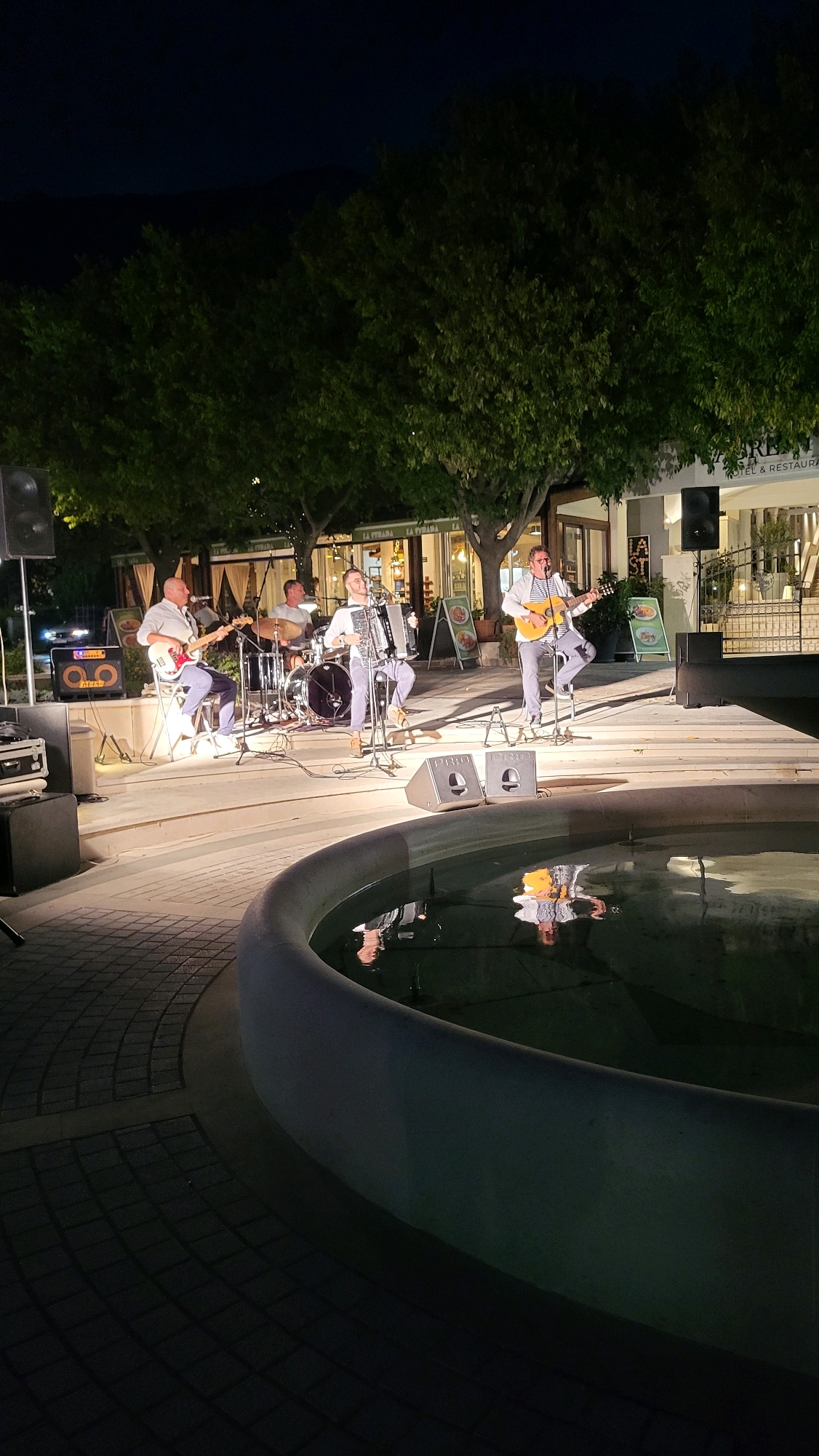 In Općina Tučepi, Croatia, at night, a band of four men performs on an outdoor stage illuminated by artificial light. The stage is set on a paved area with a decorative fountain in the foreground. The band members are positioned from left to right: a man playing a bass guitar, a man seated behind a drum kit, a man playing an accordion, and a man playing an acoustic guitar while singing. They are all dressed in light-colored shirts and darker pants. Behind the stage, trees form a backdrop, and beyond them, buildings with awnings and signage, including "HOTEL & RESTAURANT" and "LA STRADA," suggest a commercial area. Two large speakers are visible on the stage, and audio equipment is also present. The fountain's water reflects the lights and the band. The ground in front of the stage is paved with interlocking bricks.