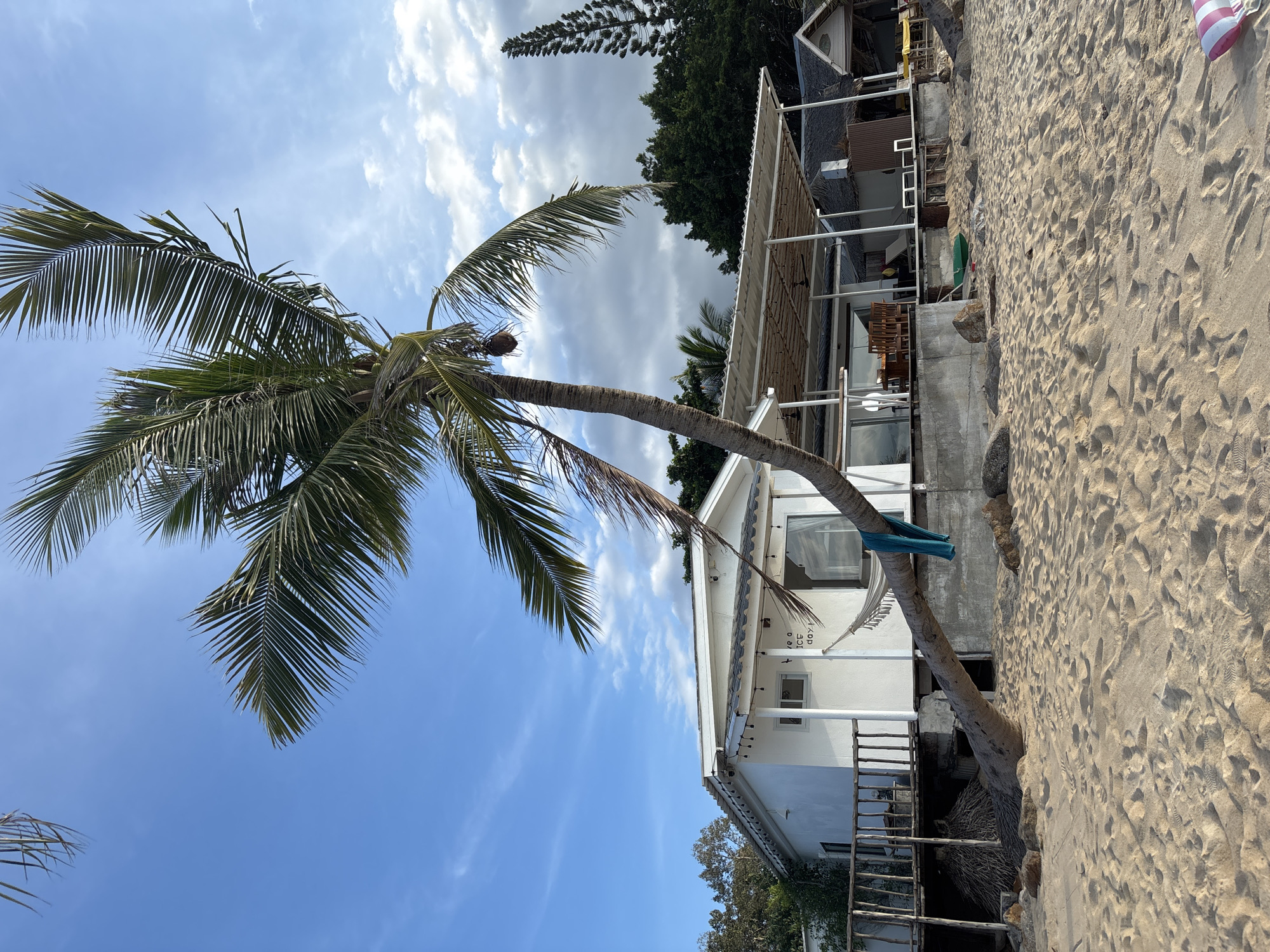 The image depicts a beach scene in Baan Mae Nam, Thailand. A tall palm tree with green fronds leans towards a white, two-story building situated on the sand. A blue fabric is tied around the trunk of the tree. The building features a covered patio area with seating. The beach is sandy with footprints visible. The sky is partly cloudy, with blue patches. Vegetation and part of another building can be seen in the background.