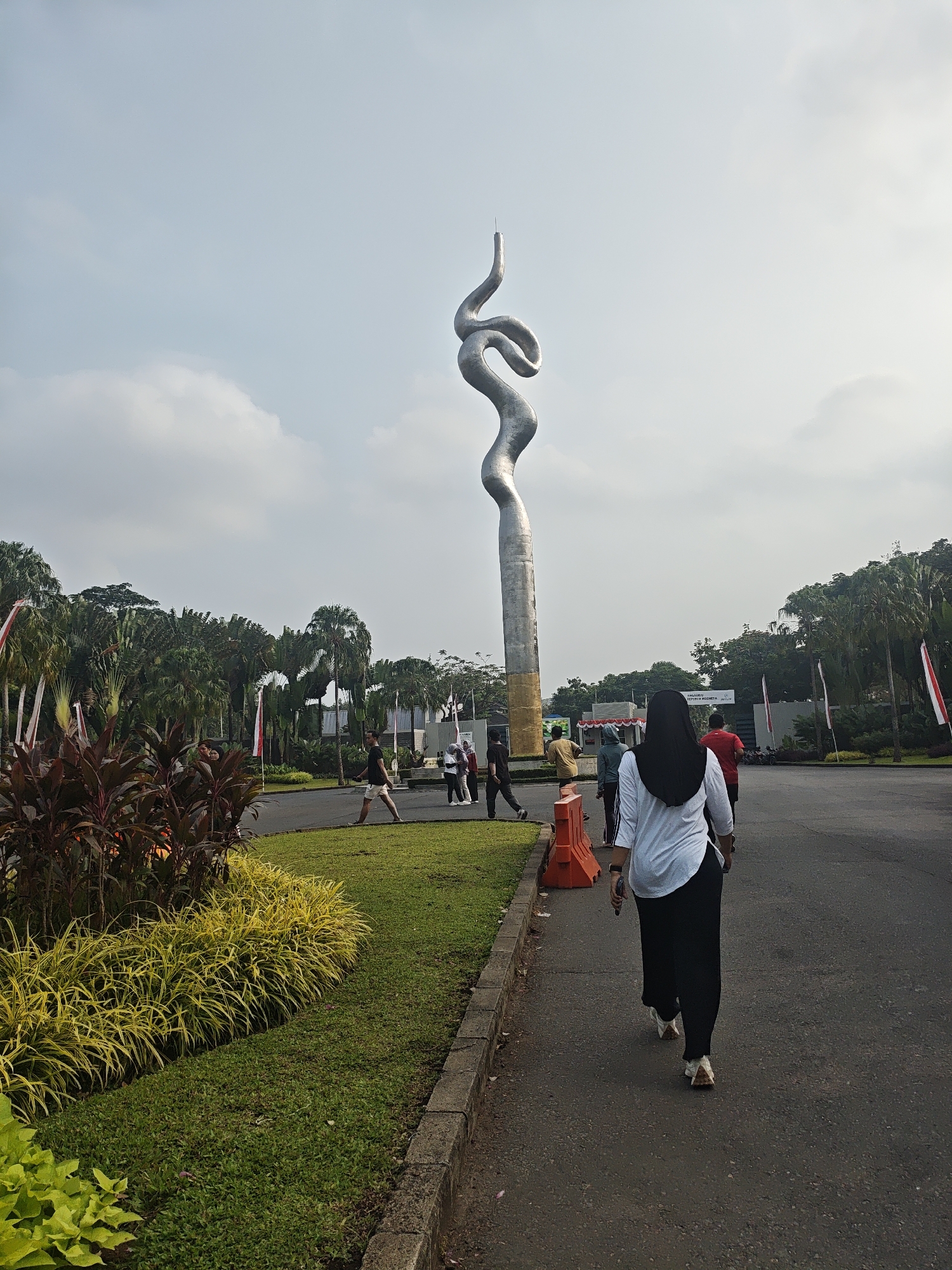 The image shows an outdoor scene with a tall, metallic sculpture resembling a twisted ribbon or snake, located in the city of Bacin, Indonesia. In the foreground, a woman wearing a black hijab and a white shirt is walking away from the camera on a paved path. To her left, a well-maintained garden features vibrant green grass, yellow ornamental plants, and dark red foliage. Behind the garden, a paved road curves to the right, with several people walking or standing in the distance. The background is filled with lush green trees, including palm trees, and a cloudy sky. Indonesian flags are visible on poles in the background. The overall scene depicts a public space, possibly a park or monument area, with people engaged in outdoor activities.