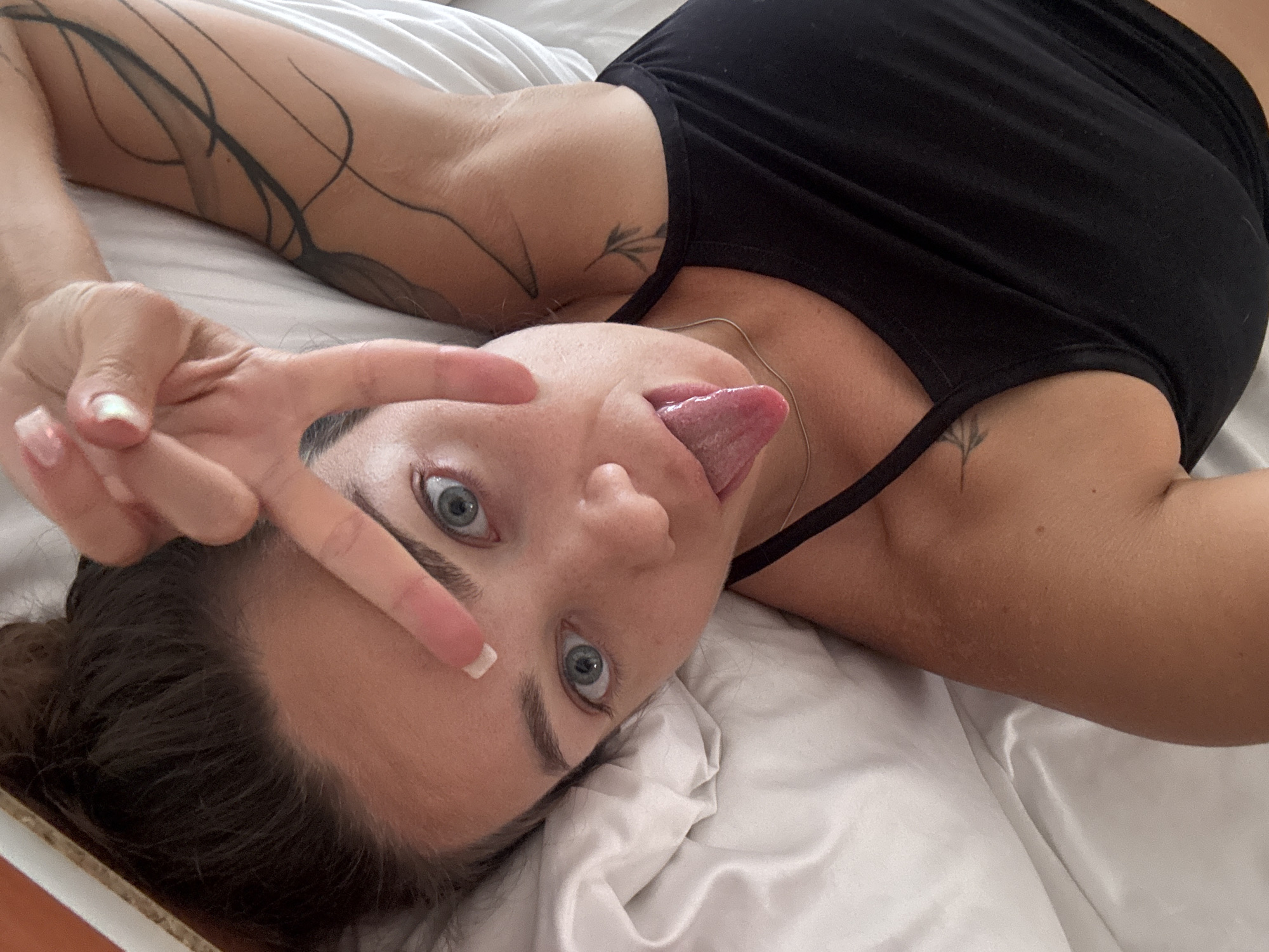 A young woman lies on a white bed, her head resting on a pillow, with her body angled towards the camera. Her eyes are wide open, looking directly at the lens, and her tongue is sticking out. She is making a peace sign with her right hand, which is held up near her forehead. A large tattoo is visible on her left arm, extending up her shoulder. She is wearing a black tank top. The scene appears to be indoors, possibly in a hotel room or bedroom. The lighting suggests it is daytime, with natural light coming from the side. The overall impression is playful and casual. The location is the city of Budva, Montenegro.