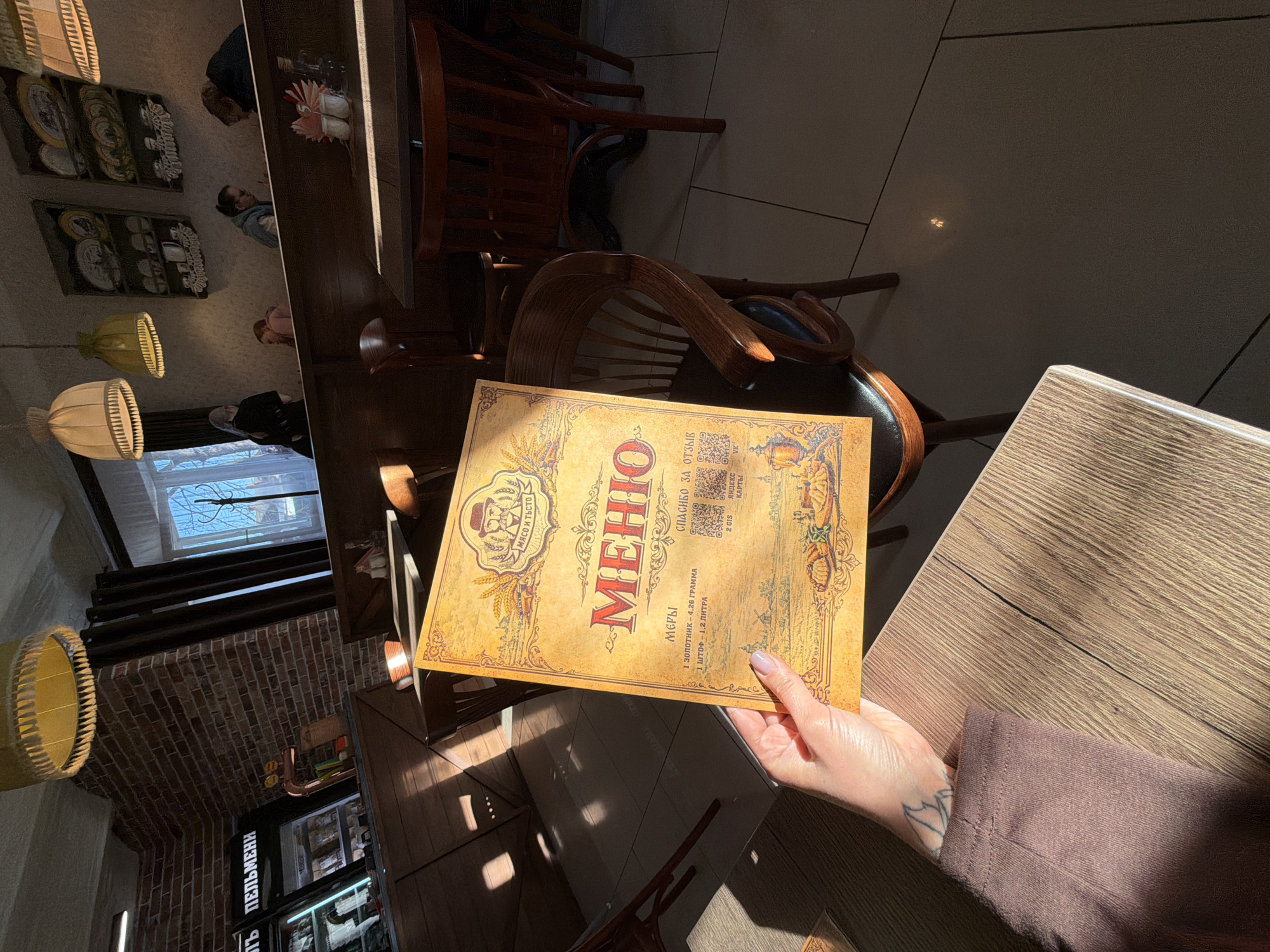 A person sits at a wooden table in a restaurant, holding a menu from "Мясо и Тесто" (Meat and Dough), which features the word "МЕНЮ" (Menu) in large letters. The menu also contains phrases like "СПАСИБО ЗА ОТЗЫВ" (Thank you for the feedback) and "МЕРЫ" (Measures), listing "1 ЗОЛОТНИК - 4,26 ГРАМΜΑ" (1 zolotnik - 4.26 grams) and "1 ШТОФ - 1,2 ЛИТРА" (1 shtof - 1.2 liters). The restaurant has wooden chairs, and a brick wall is visible in the background, along with hanging lamps. The restaurant is likely located in the city of Sarapul, Russia, as suggested by the location name and the Russian text on the menu. The person's hand with light skin and a tattoo is visible.