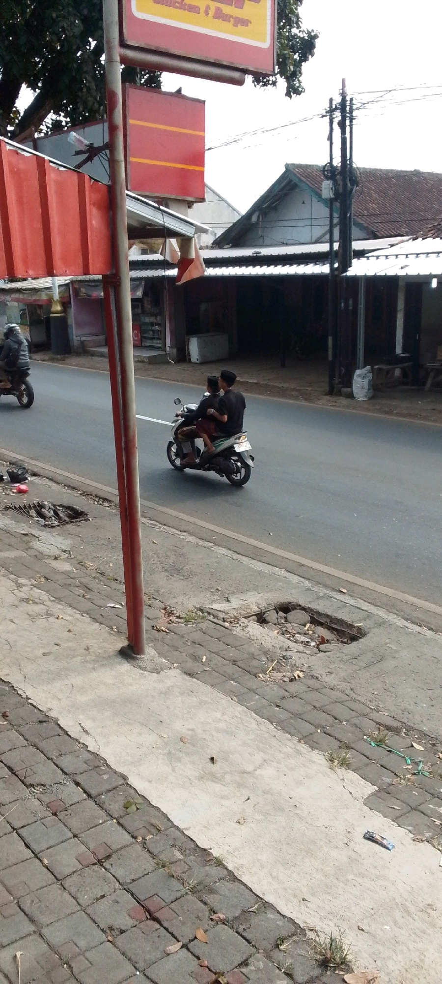 This is a street-level, eye-level, medium shot photograph taken in Cilaku, Indonesia. The image features a road with two individuals riding a motorcycle. The driver is wearing a black shirt and a black peci cap, while the passenger, positioned behind the driver, is also wearing a black shirt and a black peci cap. They are riding a white and silver scooter. To the left of the road, there is a sidewalk with cracked paving stones and a large hole. A red pole, part of a signage structure, stands prominently in the foreground. Behind the road, there are buildings with tiled roofs and an overhead power line. On the sidewalk to the left, there's a sign with red and yellow elements. The overall scene depicts a typical street in a residential or commercial area.
