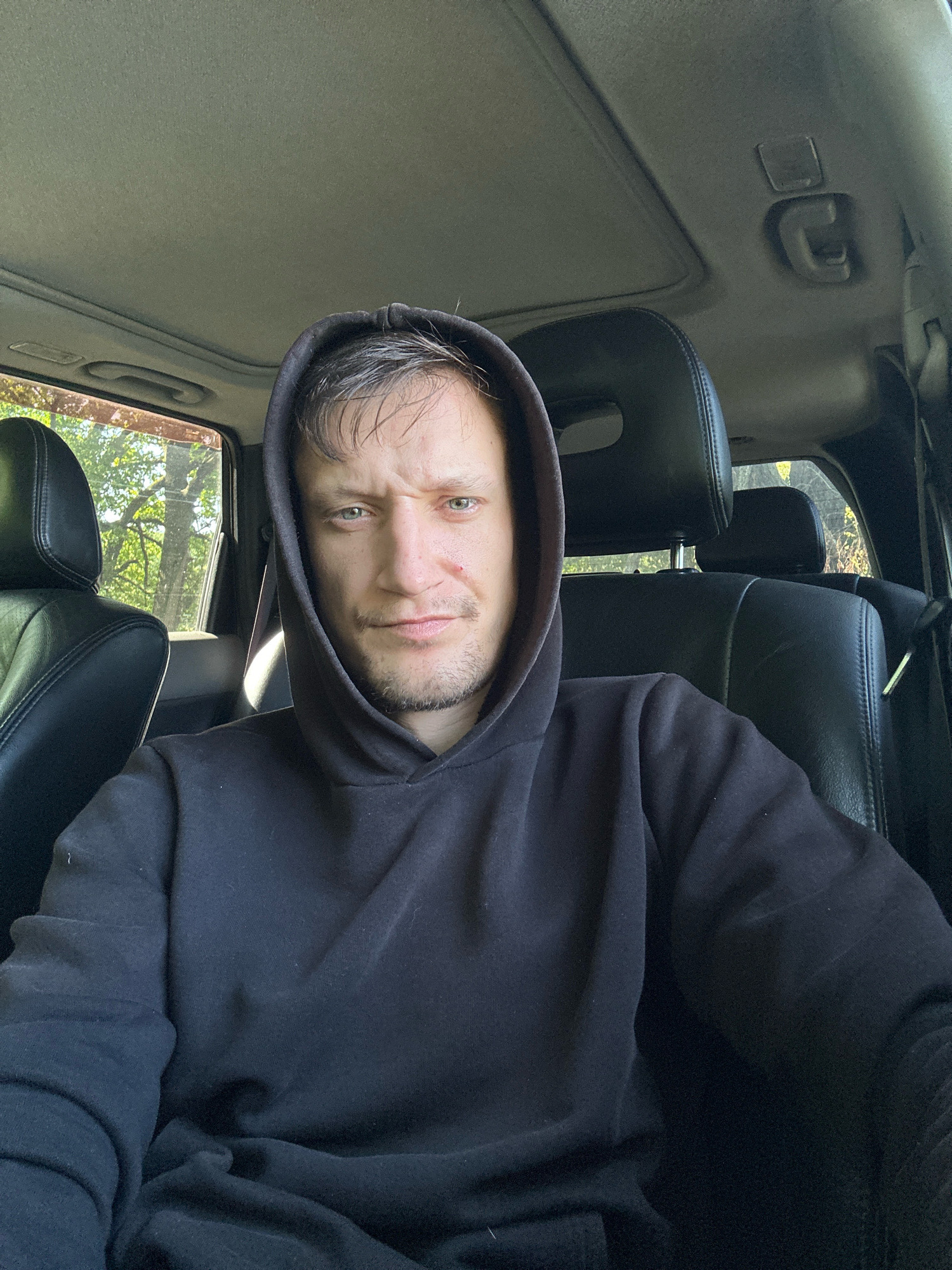 The image shows a man sitting in the driver's seat of a car. He is wearing a black hoodie and looking directly at the camera. The car's interior, including the dashboard, headrests, and a sunroof, is visible. The exterior scenery suggests a natural, possibly rural environment with trees and sunlight filtering through the windows. There is no indication of the city of Shatava, Ukraine.