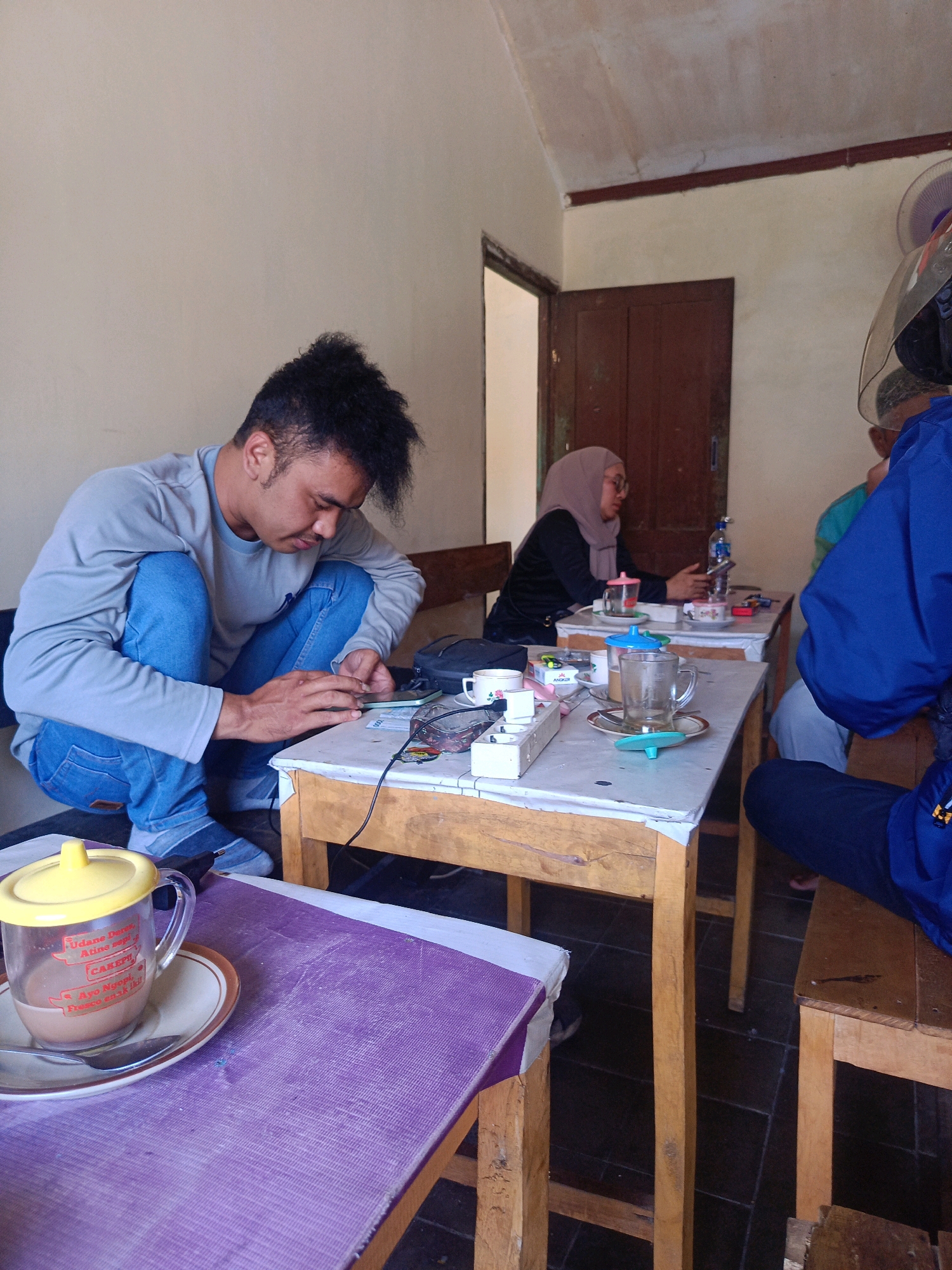 The image depicts an indoor scene at a cafe or eatery, likely in Kebonagung, Indonesia. A young man with dark, spiky hair is crouched on a wooden chair and table, intensely focused on his mobile phone. He wears a grey long-sleeved shirt and blue jeans. In the foreground, a mug with a yellow lid containing a brown beverage sits on a purple placemat, beside a spoon on a saucer. Another table in the mid-ground has various items, including cups, a bottle of water, and what appears to be a cigarette pack. Two other individuals are partially visible at this table: a woman wearing a light brown hijab and a black top, and the back of someone in a blue jacket, possibly another customer or staff member. The environment appears modest, with simple wooden furniture and light-colored walls. The flooring is dark tile. A partially open wooden door is visible in the background, leading to an unknown area. The lighting suggests it is daytime.