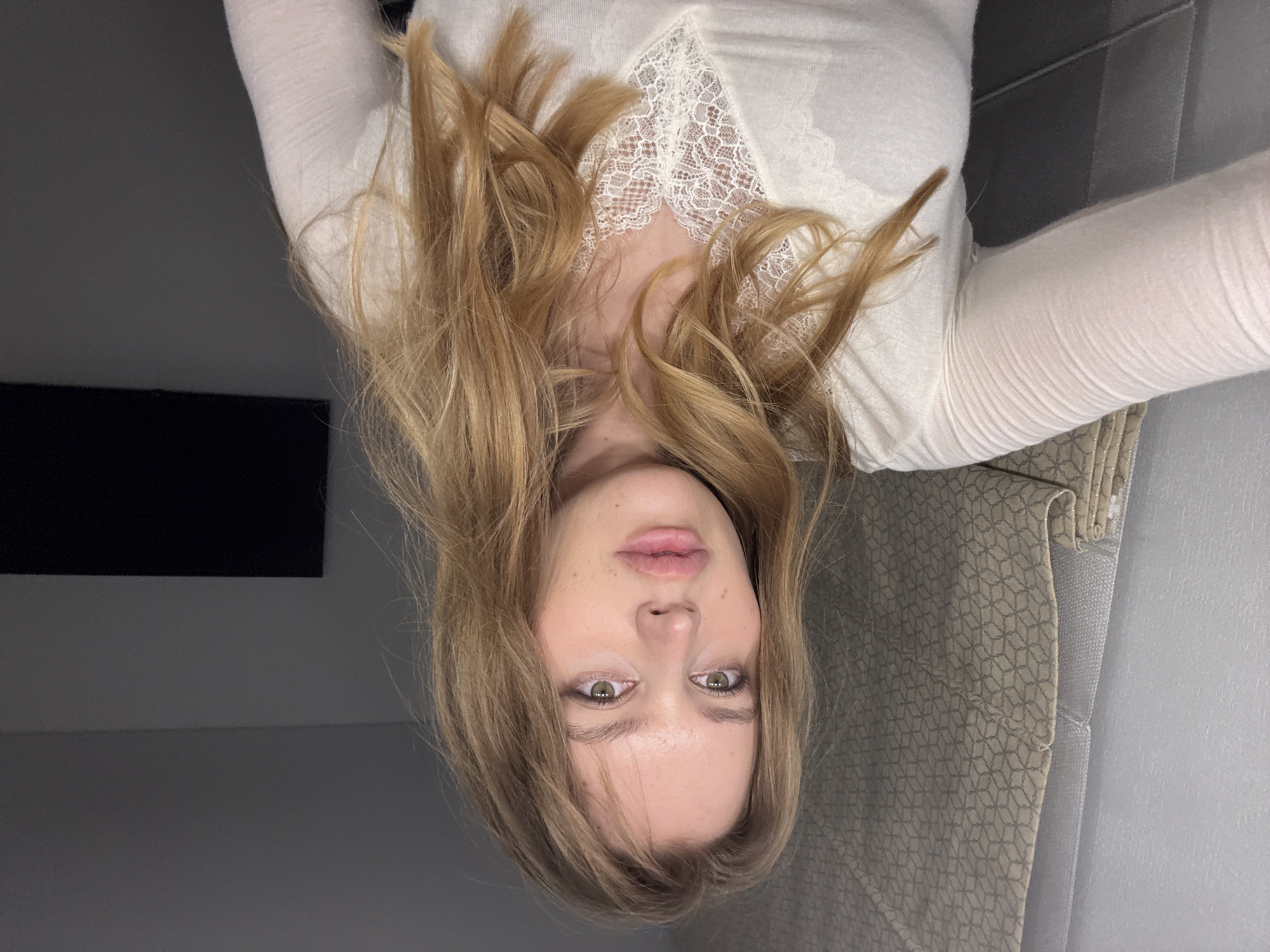 A photograph depicts a woman with fair skin and light brown hair, positioned upside down in the frame. She is wearing a white, long-sleeved garment with lace detailing at the neckline. Her eyes are light-colored, and she has full lips. The background consists of a gray wall with a dark, rectangular object, possibly a television, mounted on it. A patterned window covering is visible near the bottom right of the frame. The location is not specified. The picture does not give the location in the city of Hrodna, Belarus.