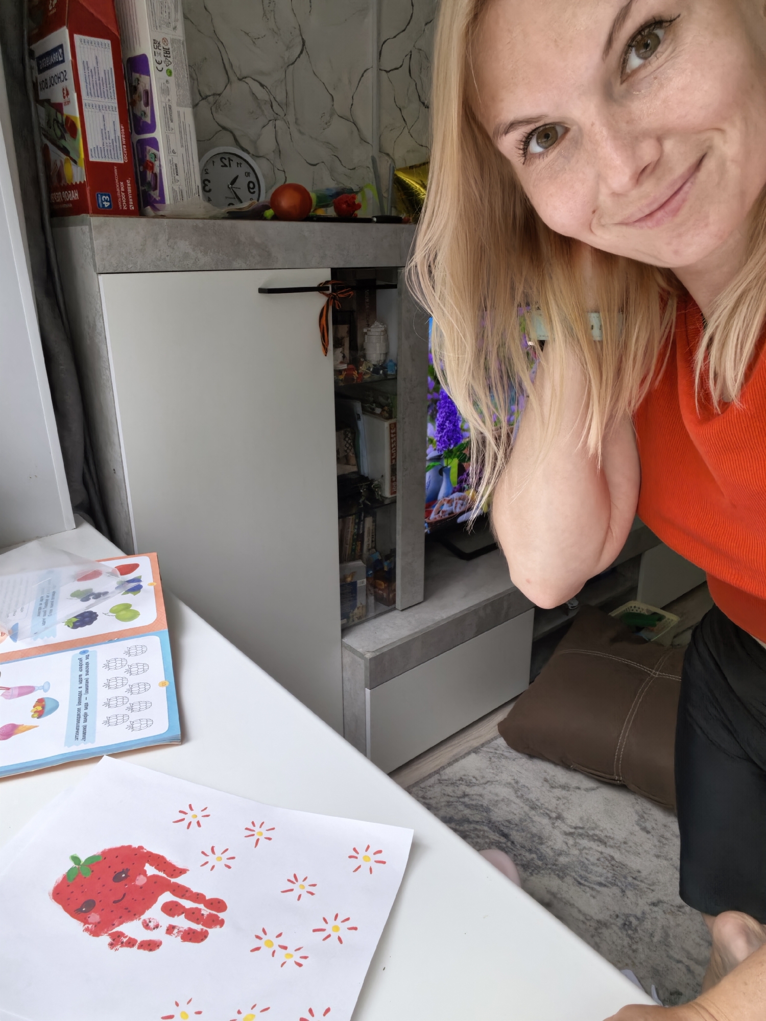 The image shows a woman with blonde hair and a friendly smile looking at the camera. She is wearing an orange shirt. In the background, there is a white cabinet with a shelf above it containing various items including boxes, a clock, and decorative objects. To the left of the woman, a table holds an open book with colorful illustrations of fruits and ice cream, and a piece of paper with a handprint strawberry artwork and small yellow flower drawings. The room appears to be a domestic setting, possibly a living room or playroom. The image does not contain any specific information that would indicate its location in Sterlitamak, Russia.