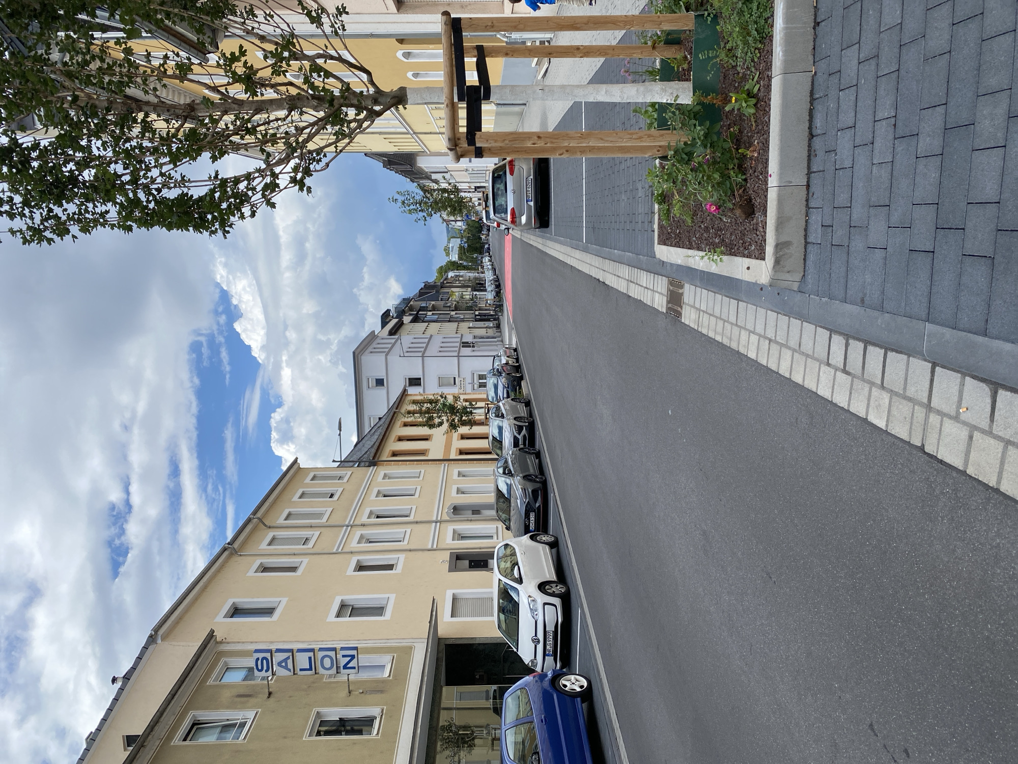 The image depicts a street scene in Neuwied, Germany. On the left side of the street, a yellow building with a blue sign reading "SALON" vertically mounted on its facade is visible. Several parked cars, including a blue hatchback and a white compact car with license plate "NR. G 9797", are lined up along the curb. The street is paved with asphalt, and there is a distinct red-painted section further down the road, possibly indicating a bike lane or a special traffic zone. On the right side of the street, a young boy in a blue shirt and shorts is walking away from the camera. A newly planted tree is secured with wooden stakes and dark straps in a planter box. The surroundings are characterized by multi-story buildings and a cloudy sky.