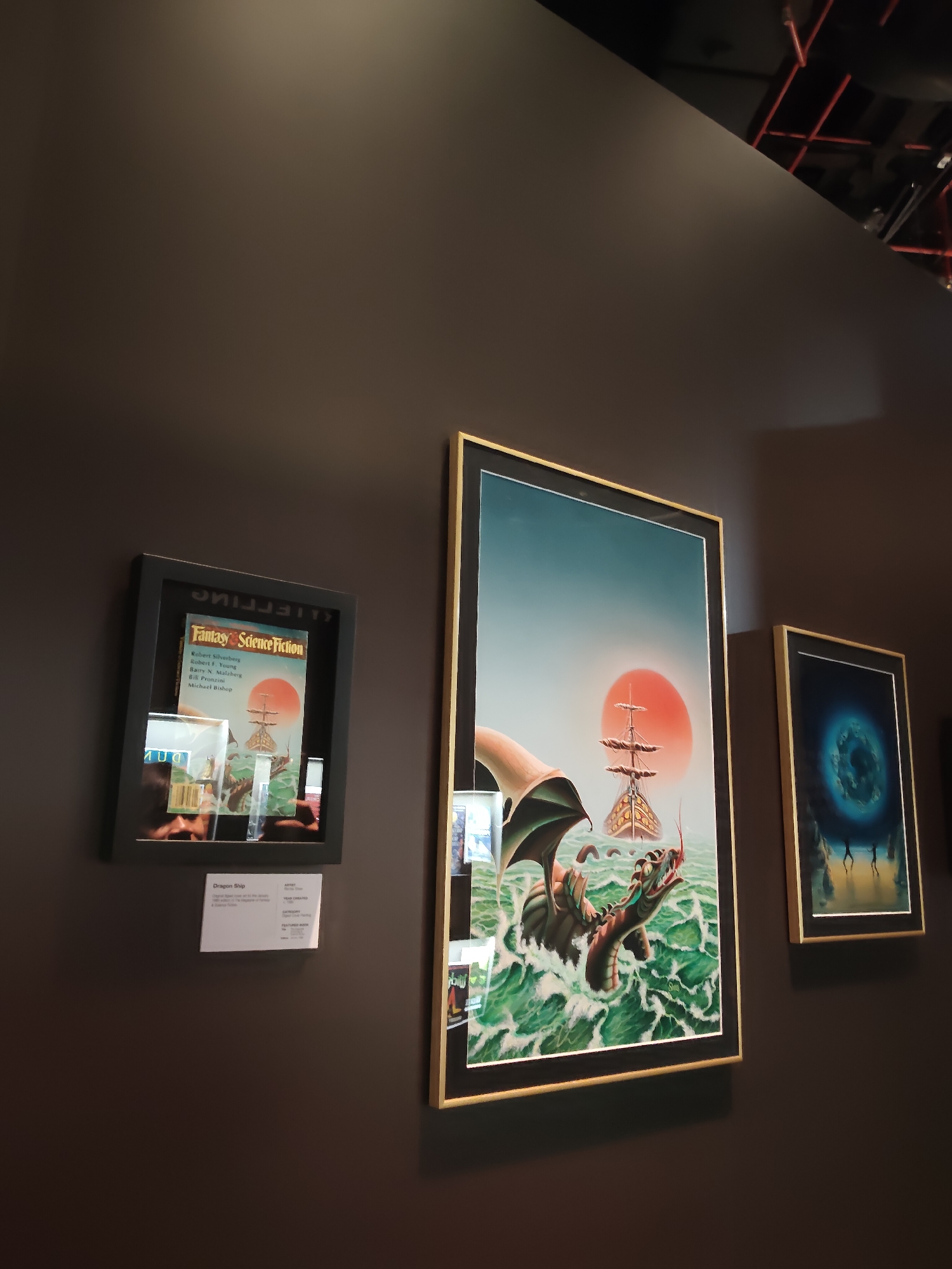 The image shows a museum exhibit with three framed pieces on a dark brown wall. On the left, a black frame displays a vintage magazine cover titled "Fantasy Science Fiction" with an illustration of a dragon and a sailing ship in stormy seas. Below the magazine, a small information card reads "Dragon Ship" and describes the artwork. To the right of the magazine display are two larger framed art pieces. The central piece is a framed illustration of a dragon attacking a sailing ship with a large orange sun in the background, set against a gradient sky. The rightmost framed piece depicts a surreal, swirling blue and green vortex with figures standing on a dark shore. The lighting suggests an indoor setting, likely a gallery or museum, with a glimpse of exposed ceiling infrastructure with red lighting in the upper right corner. The overall scene is static, showcasing a collection of art. No specific location in Singapore is identifiable from the image.