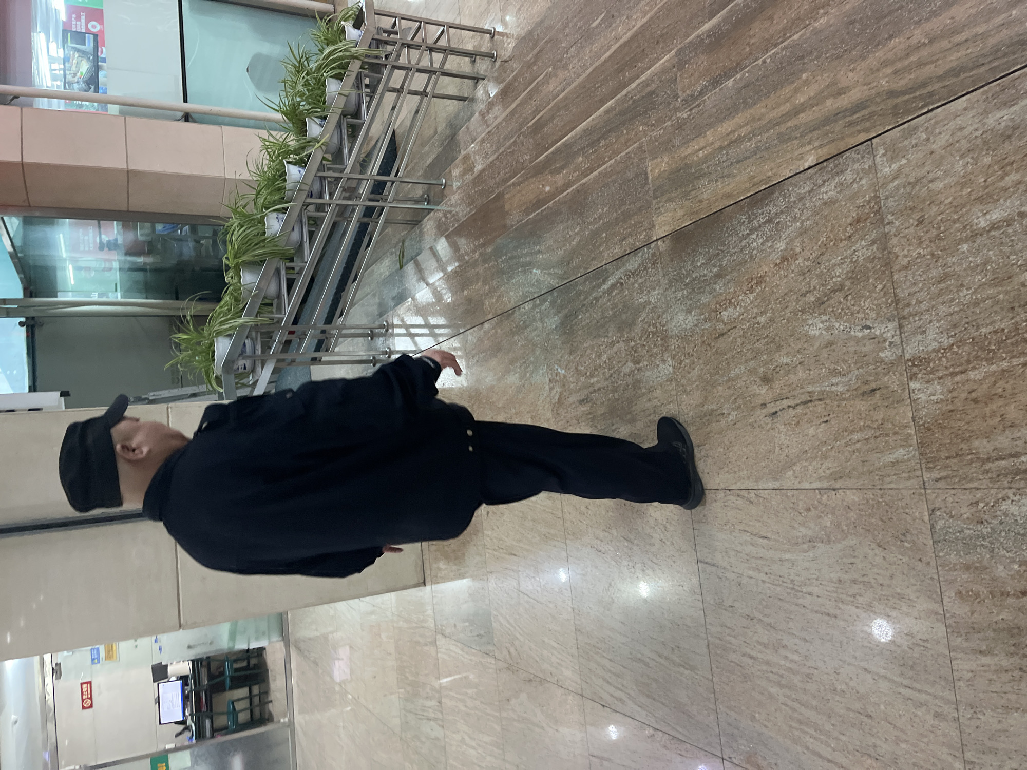 A medium, rotated, indoor shot shows a person walking on a light beige tiled floor. The person is wearing a black cap, a black jacket, and black pants and shoes. To the left of the person is a partially-obscured glass wall, some plants on a metal shelving unit, and some electronic equipment. To the right of the person is a tan tiled area with multiple steps and metal railings. The location is in Changchun City, China.