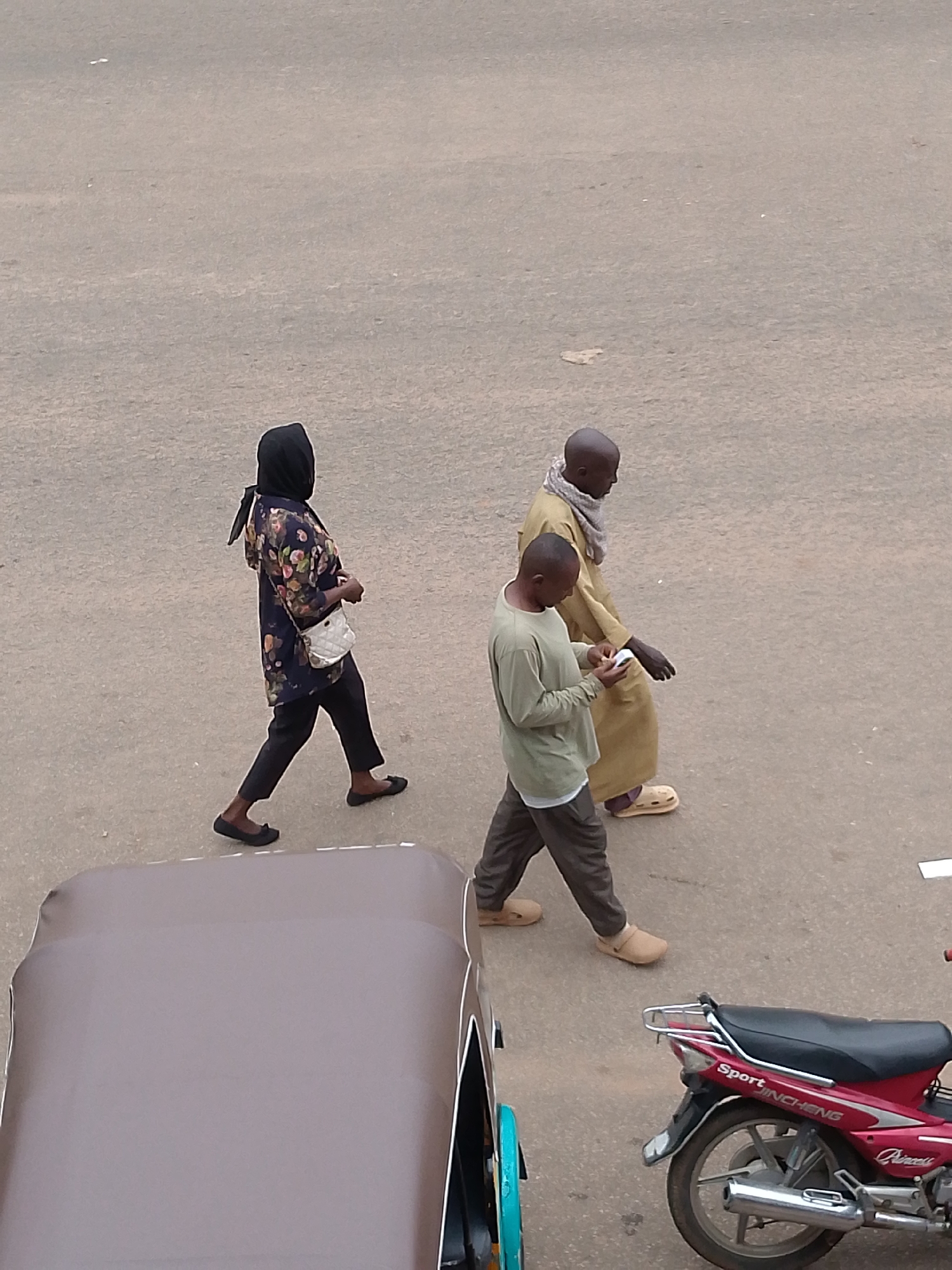 In Bukuru, Nigeria, three individuals are walking on a paved road. A woman in the foreground, wearing a black hijab and a floral print tunic over black trousers and black flats, carries a white crossbody bag. To her right, a man in a long, light brown robe and a scarf walks with a younger man wearing a light green long-sleeved shirt and dark trousers. The younger man is looking at his phone. Partially visible in the bottom left is the canopy of a motorized tricycle, and to the right, a red motorcycle is parked. The environment appears to be an outdoor urban setting.