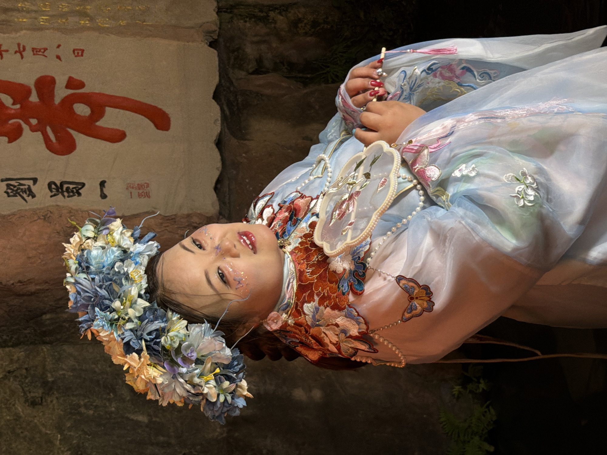 A young woman is depicted in a close-up, horizontally oriented shot, wearing a blue and white flowing garment adorned with intricate floral embroidery and beaded chains. She has a crown of flowers on her head and glitter makeup on her face, giving her a whimsical appearance. Her hands, with manicured red nails, are delicately holding a small, ornate fan. The background appears to be a stone wall, suggesting an outdoor or ancient setting, possibly in Yuzhong District, China. A stone tablet with red calligraphy is visible in the upper left corner of the image. The lighting suggests it might be evening or a dimly lit indoor space. The woman's expression is serene, with her eyes looking towards the viewer.