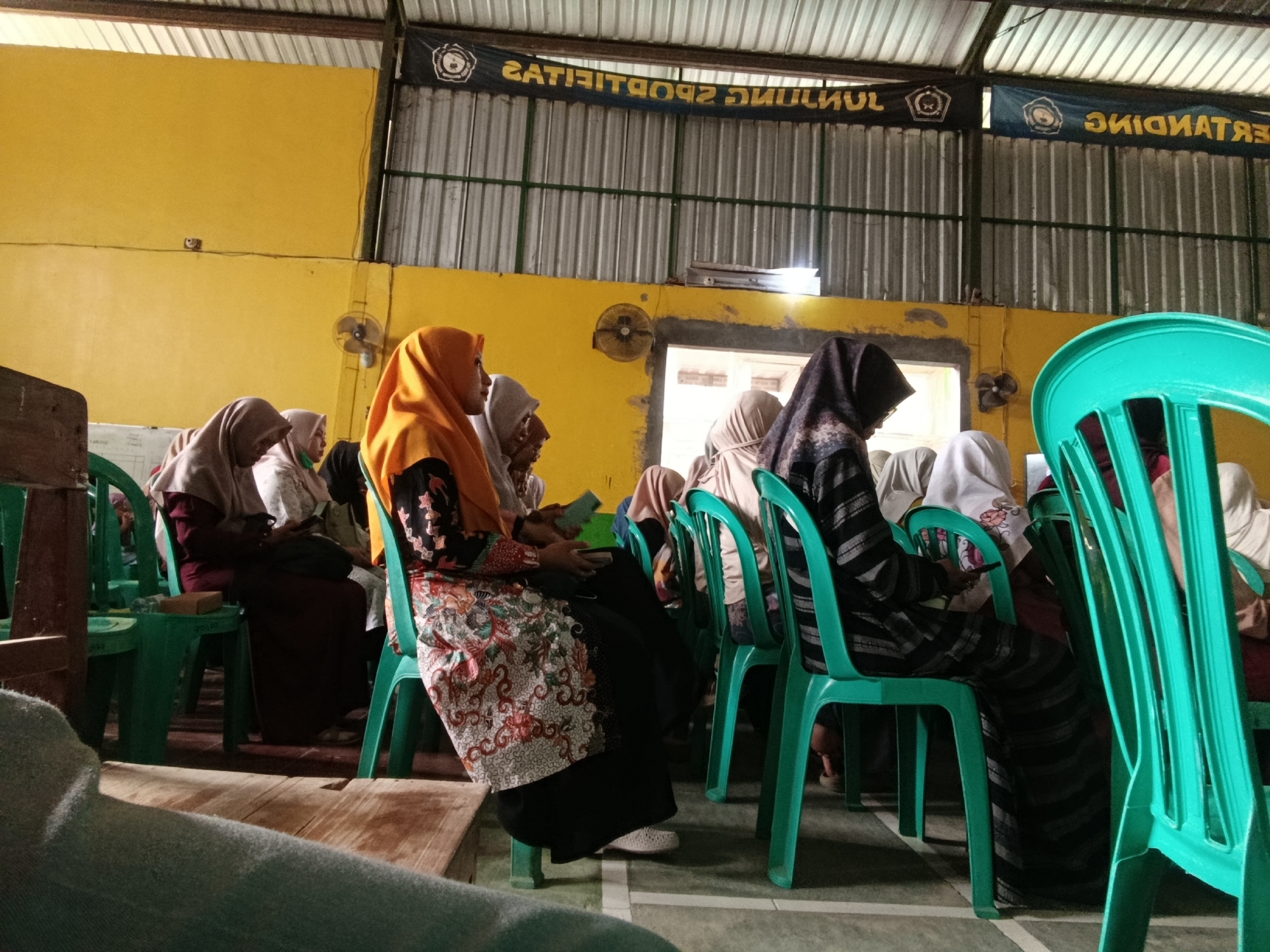 The image depicts a group of women wearing hijabs, seated on teal plastic chairs in what appears to be an indoor community hall. The hall has a yellow wall and a corrugated metal ceiling. There are fans mounted on the wall. Banners with text and logos are visible hanging from the ceiling. The women are facing forward, and some are holding phones. One woman in the foreground is wearing a patterned skirt and an orange hijab. The flooring is a dark, possibly tiled, surface with white lines marking sections. The scene is likely within a public building in Kawunganten Lor, Indonesia, potentially during a gathering or event.