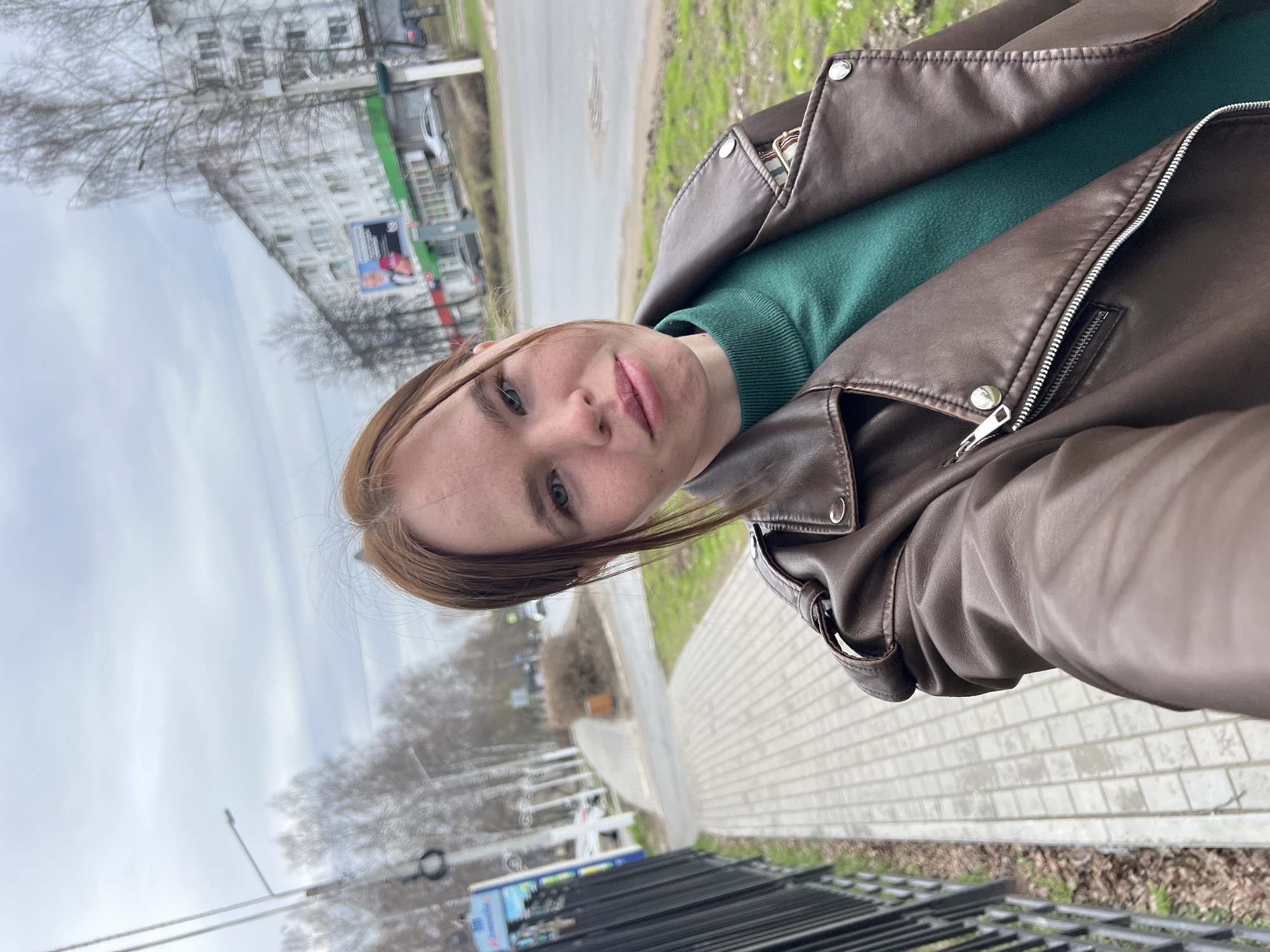 A woman is taking a selfie outdoors. She is wearing a brown leather jacket over a green sweater. The background includes a sidewalk, a street with some traffic, a green strip of grass, trees with bare branches, and a building. The photo was taken on a cloudy day. While a precise determination of the location in Chaikovsky, Russia, is not possible, the general appearance aligns with an urban setting.