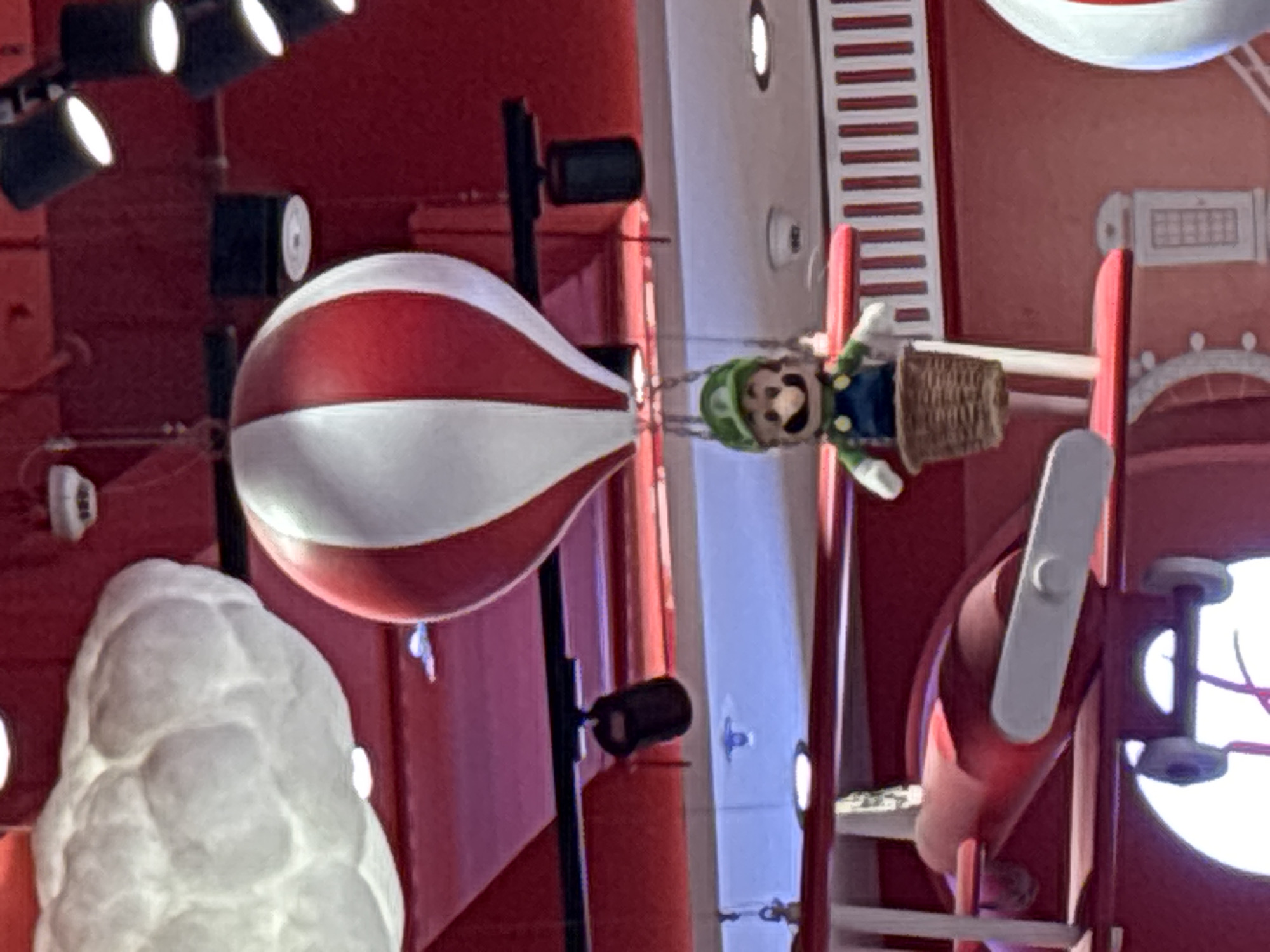 A Luigi doll, dressed in his signature green cap and blue overalls, is suspended in a wicker basket beneath a red and white striped hot air balloon. The balloon and doll are part of a decorative display, likely in a retail or entertainment venue. To the left of the balloon, a red toy airplane is partially visible, suggesting a playful, themed environment. The background consists of red walls and ceiling with industrial lighting fixtures. This display is located at the Nintendo store in Dubai, United Arab Emirates.