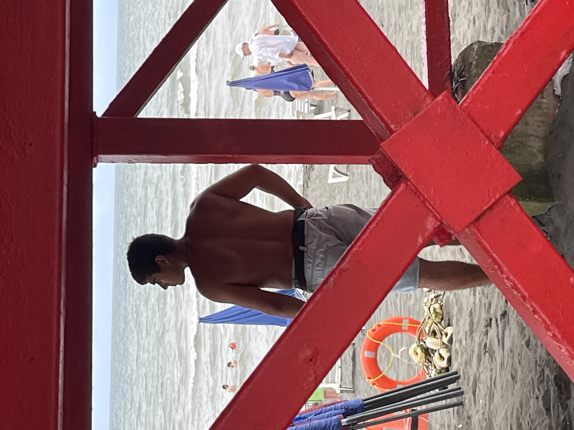 A man with dark hair is standing on a sandy beach, facing the ocean. He is shirtless and wearing light-colored shorts and a belt. The man is standing under a red metal structure, possibly a lifeguard stand or a part of a beach shelter. In the background, the ocean is visible with gentle waves and a cloudy sky. There are several beach umbrellas, some closed and some open, and people wading in the water. A lifebuoy and some buoys are on the sand near the man. The scene appears to be a typical day at the beach in Ureki, Georgia, a coastal city known for its magnetic sand beaches.