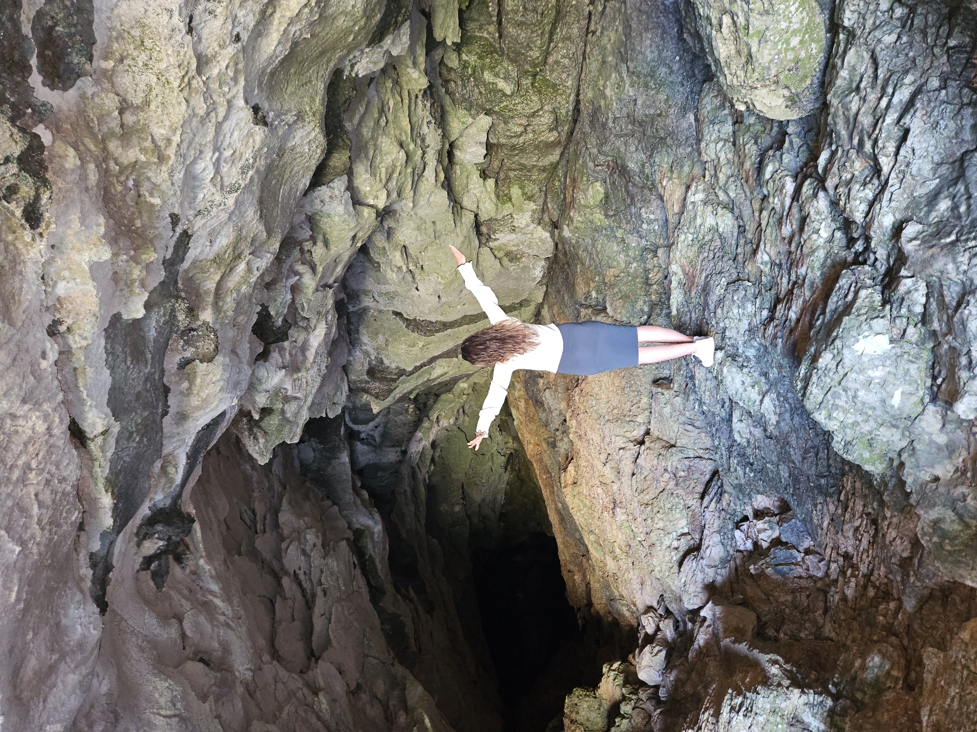 A woman is seen from the back standing inside a cave. She is wearing a white shirt and a blue skirt. Her arms are extended outwards. The cave walls are rough and textured, with varying shades of gray, brown, and green. The setting appears to be dimly lit. The location is Unknown, Unknown.