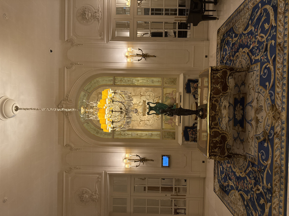 A grand hall features an ornate chandelier, a gilded sculpture, and a patterned rug. Architectural details include arched windows with decorative paintings, wall sconces, and intricate moldings. A table sits atop the rug, positioned beneath the sculpture. The room exudes a sense of opulence and classic elegance.