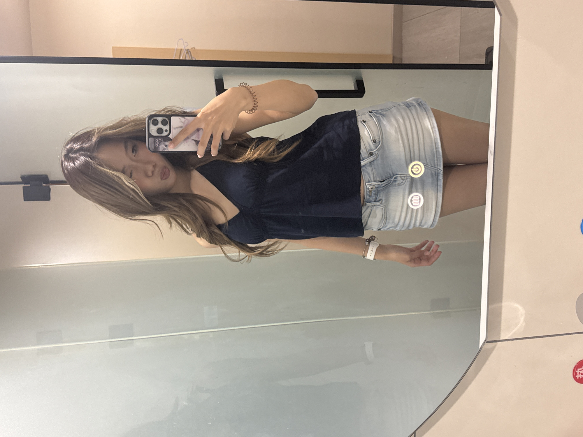 A young woman is taking a mirror selfie indoors, likely in a modern fitting room or bathroom. The image is oriented with the woman's head to the left, standing vertically in the reflection. She has long, light brown hair and wears a dark blue sleeveless top and a light-wash denim mini-skirt. Her facial expression is playful, featuring a slight pout and a partially closed eye.
She holds a smartphone with a light-colored, marble-patterned case to capture her reflection. On her left wrist, she wears a spiraled metallic bracelet, and on her right, a white smart band or watch. The mirror itself appears to be a smart mirror, displaying illuminated circular icons: a yellow/gold power symbol and white wavy lines (possibly indicating a heating or fan function) reflected over her skirt.
The background reflected in the mirror includes a light-colored wall and a shelf with clothes hangers. To the right of the mirror, outside the reflection, a light-colored wall displays red and blue square symbols with white Chinese characters. The red symbol appears to be "折" (zhé), commonly associated with "discount" or "promotion," suggesting a retail environment. The overall setting is clean and modern, typical of urban interiors in Yuzhong District, China.