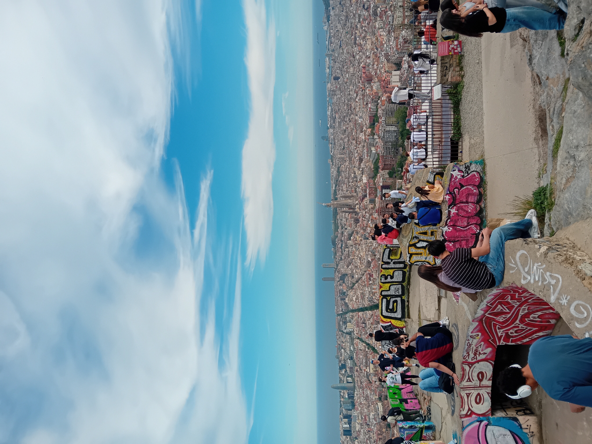 The image shows an elevated view of the city of Barcelona, Spain. A crowd of people is seen sitting and standing along a graffiti-covered ledge, overlooking the cityscape and the Mediterranean Sea. The sky is a mix of blue and white clouds. The architecture of Barcelona, including tall buildings and landmarks, is visible. The people are casually dressed, and some appear to be taking pictures or enjoying the view. The ledge is concrete and adorned with a variety of colorful graffiti.