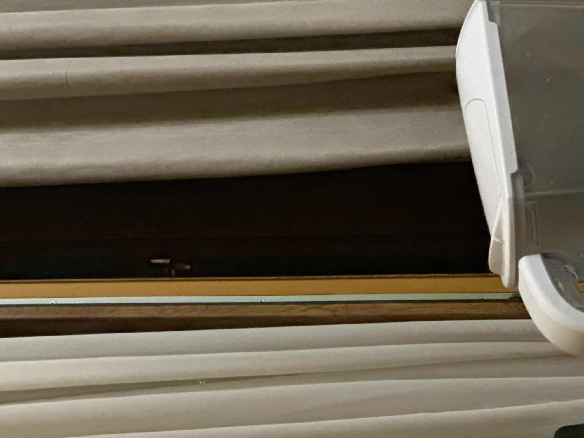 The image shows a close-up view of window blinds, partially open. The blinds are light gray or beige in color. A white object, possibly part of the blind mechanism or a separate device, is attached to the right side of the window frame. The window frame appears to be made of wood, with a dark stain. There is a dark gap between the opened blinds. The environment is otherwise undiscernible. The city location is not discernible.