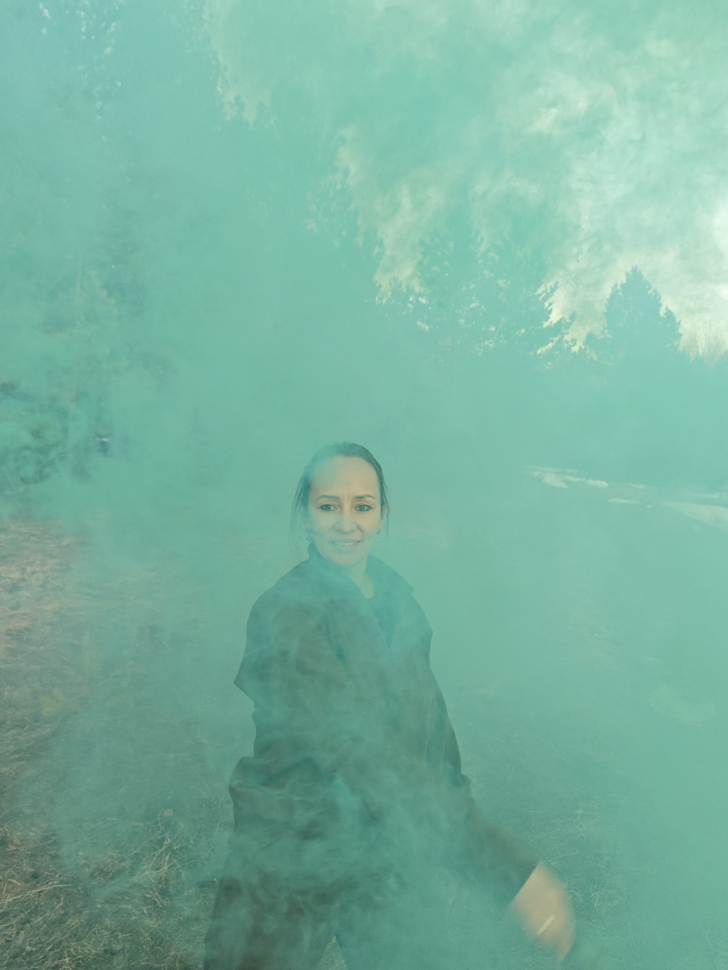 A medium-shot portrait captures a woman standing amidst dense, vibrant teal-green smoke in an outdoor setting in Dubovaya, Russia. The woman, positioned centrally, looks directly at the viewer with a slight, warm smile. Her dark hair is pulled back, and she is wearing a dark jacket or coat.
The overwhelming presence of the teal smoke dominates the scene, creating an ethereal and hazy atmosphere. Through the thick smoke, silhouettes of trees are faintly visible in the upper background, suggesting a wooded or natural area. The ground below the woman, partially obscured, appears to be covered in dry grass or earth. The lighting is diffused, consistent with daylight filtered through the colored smoke. No text is visible in the image. The overall impression is one of a stylized photoshoot or a playful moment involving a smoke bomb.