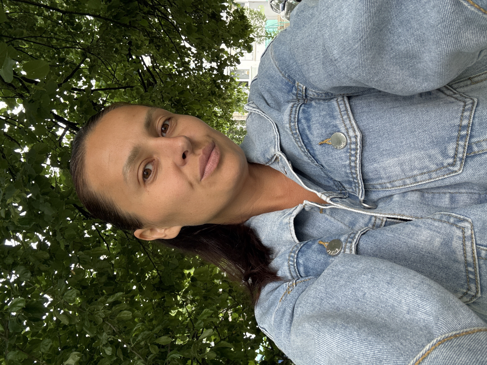 A woman is pictured from a low angle, against a backdrop of trees and a distant building. She is wearing a light-blue denim jacket. The location is purportedly Chaikovsky, Russia, though this cannot be independently verified from the image.