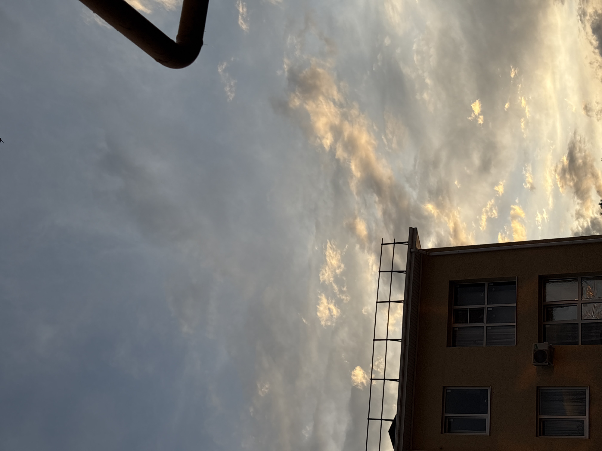 The image shows a building against a cloudy sky. The building appears to be made of a light-colored material and has several windows. There is scaffolding on the side of the building, and an air conditioning unit is mounted on the wall. In the upper left corner, a large dark pipe is visible. The sky is filled with clouds, some of which are illuminated by what appears to be the setting sun, casting a warm glow. The overall scene suggests an urban or suburban setting in Mikhaylovsk, Russia, likely during late afternoon or early evening due to the lighting conditions. There are no people visible in the image.