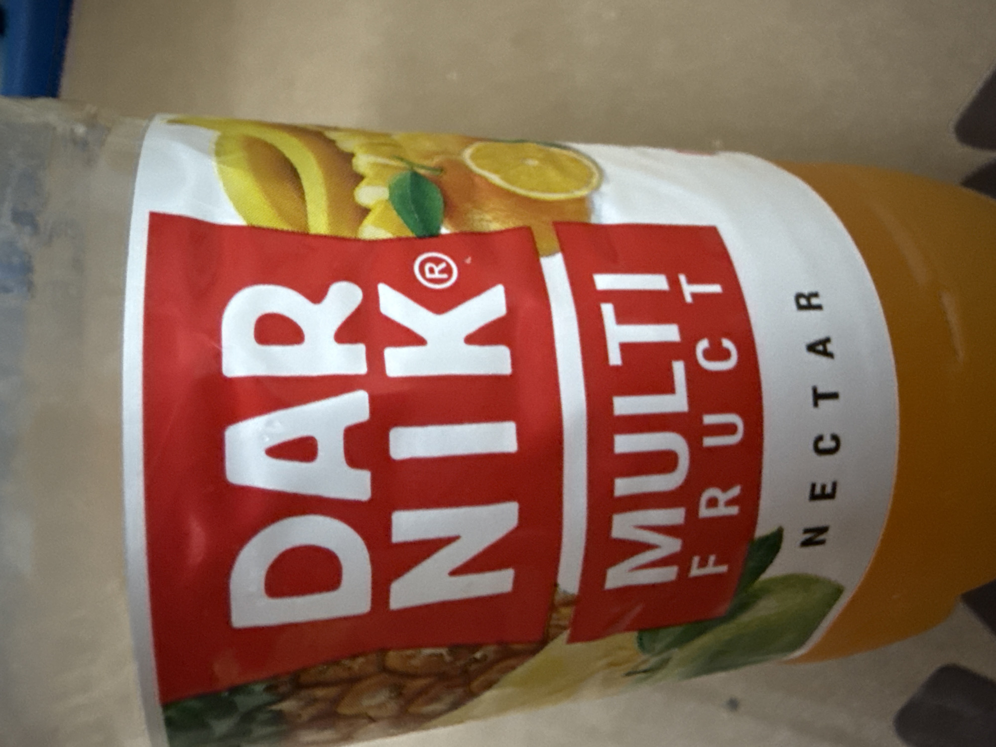 A partially visible plastic bottle of "DARNIK MULTI FRUCT NECTAR" juice is shown. The label is predominantly red and white with images of fruit such as bananas, oranges, and pineapples. The bottle contains an orange-colored liquid. The background is out of focus and appears to be an indoor setting, possibly a kitchen or pantry, with a neutral-colored wall. There are no people visible in the image. The lighting is natural and appears to be coming from the side. The image is taken at a slight angle, showing the side of the bottle. The city of Cahul, Moldova, is not depicted or implied in this image.