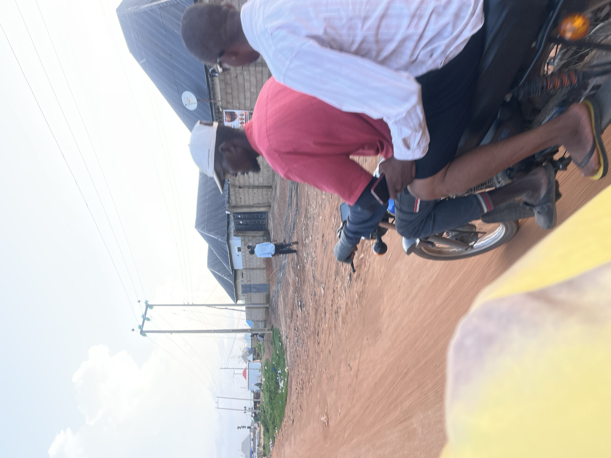 Two people are riding a motorcycle on a dirt road. The person in front wears a red shirt and a white cap. The person behind is wearing a white shirt. The background contains buildings and power lines. The setting appears to be in a city, possibly Abuja, Nigeria.