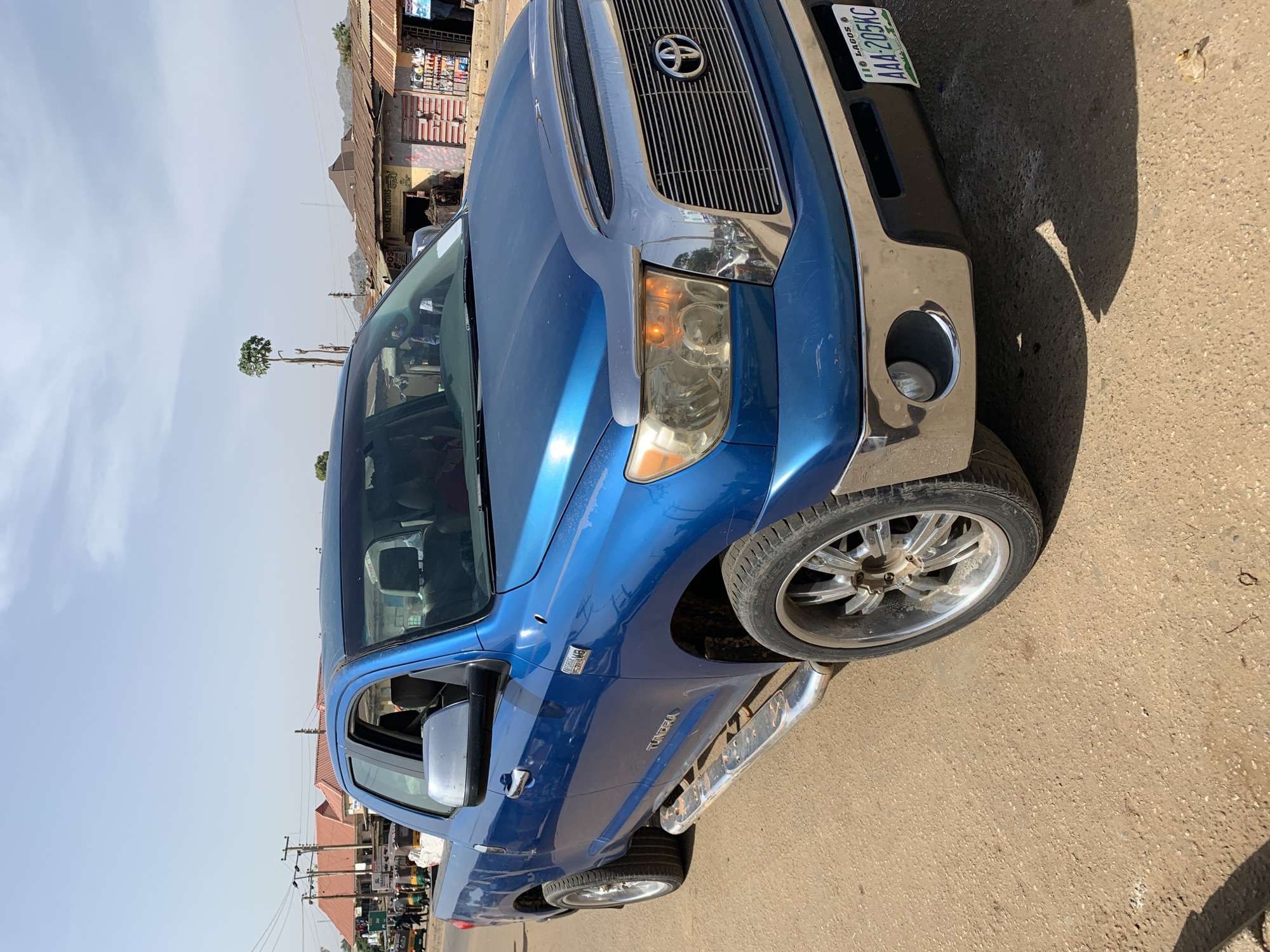 The image shows a blue Toyota Tundra pickup truck parked on the side of a road in what appears to be Rafin Par, Nigeria. The truck is angled towards the viewer, with its front end and driver's side door visible. The license plate reads "AAA-205KC" and indicates registration in Lagos, Nigeria. The immediate environment is an asphalt road. In the background, there are buildings with corrugated metal roofs and signs indicating commercial activity. Power lines and poles are also visible in the background, further suggesting an urban or semi-urban environment. There are no people visible in the immediate foreground. The light suggests it is daytime.