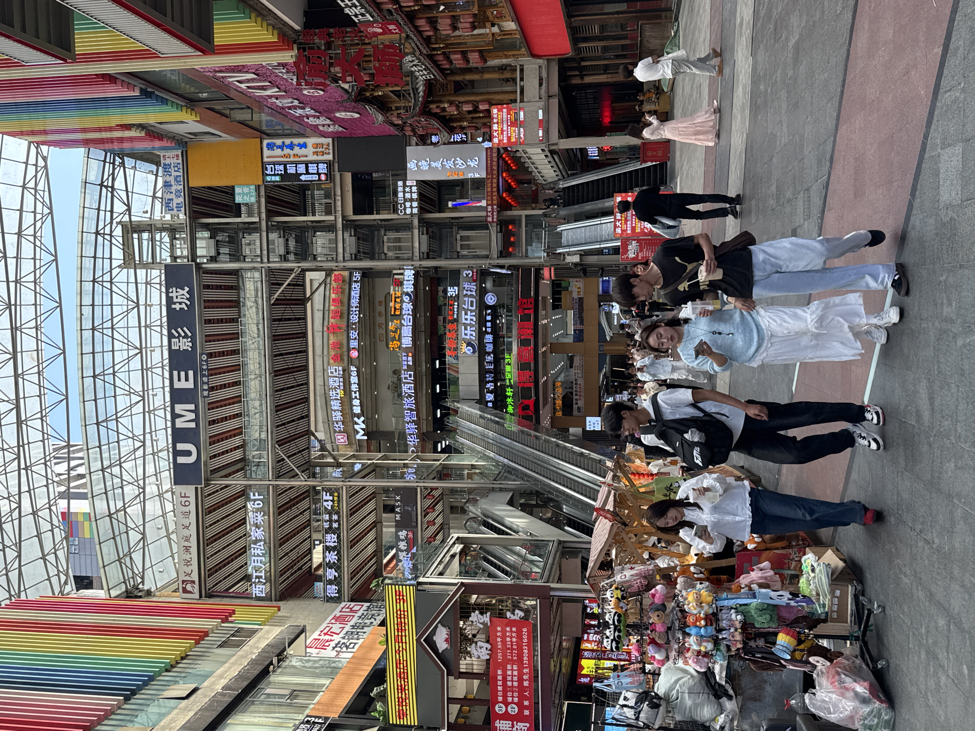 The image shows a bustling commercial street in Yuzhong District, China. Tall buildings line the street, covered in a dense array of signs in Chinese characters, including those for restaurants, hotels, cinemas (UME影城), and other businesses. Several escalators are visible within the structures, indicating multi-level shopping areas. A metal frame structure covers part of the street, with colorful rainbow-striped beams beneath. People are walking along the paved street, including couples and small groups. Some are dressed in casual attire, while others are in more formal clothing. A vendor displays various items on a cart, including stuffed toys. The lighting suggests it is daytime.