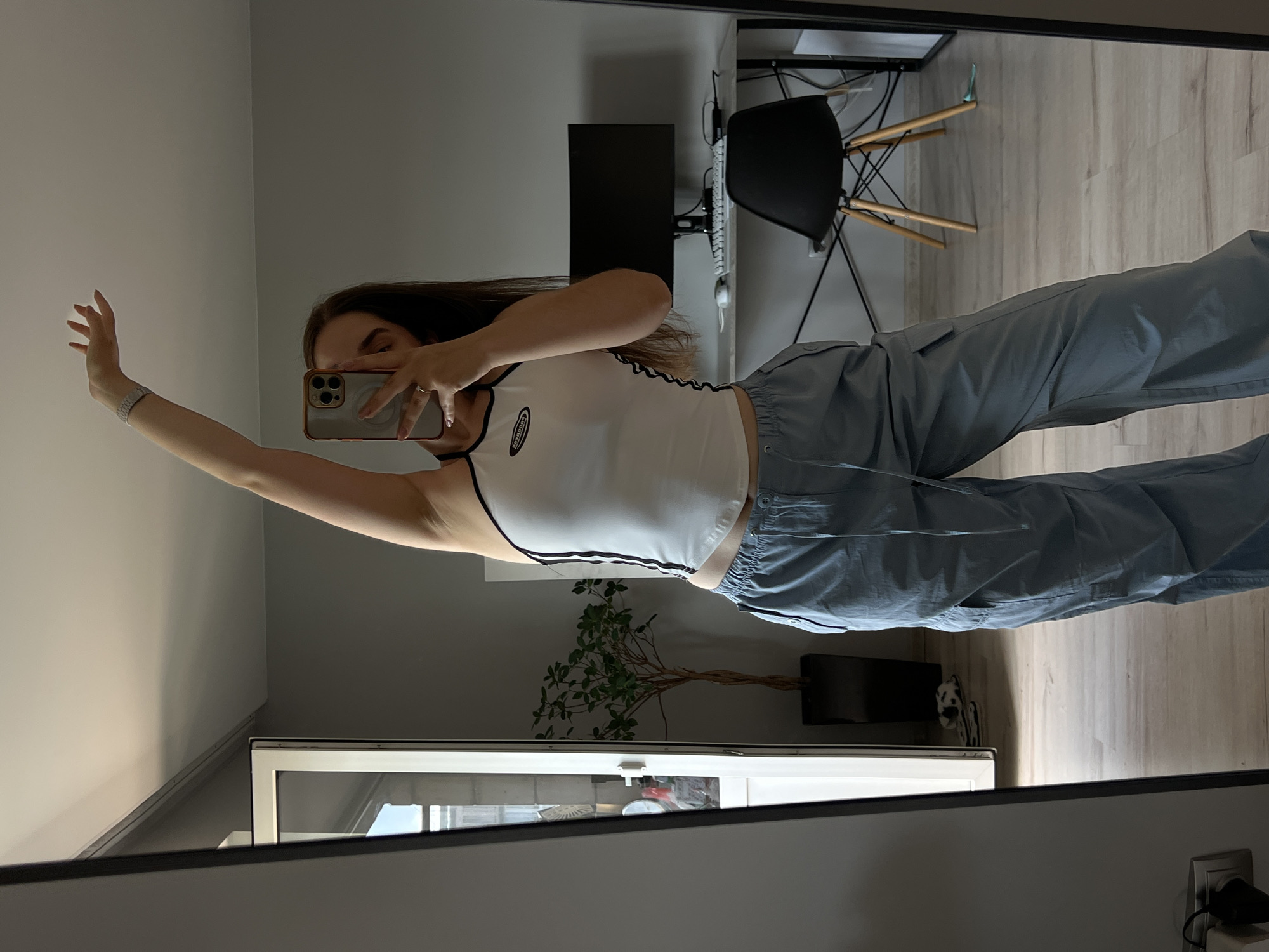 The image shows a person taking a mirror selfie in what appears to be a bedroom or a home office. The person, a young woman with dark hair, is wearing a white tank top with black trim and light blue pants. She is holding a smartphone in her right hand, capturing the reflection. Her left arm is raised, and her hand is open.
In the background, a desk with a computer monitor and a chair are visible, suggesting a workspace. There is also a plant in a pot. The room has light-colored walls and a wooden floor. The overall lighting suggests it might be daytime, but the room is somewhat dimly lit. The presence of a mirror indicates the photo is taken indoors, likely in a residential setting. There is no visible text in the image.
