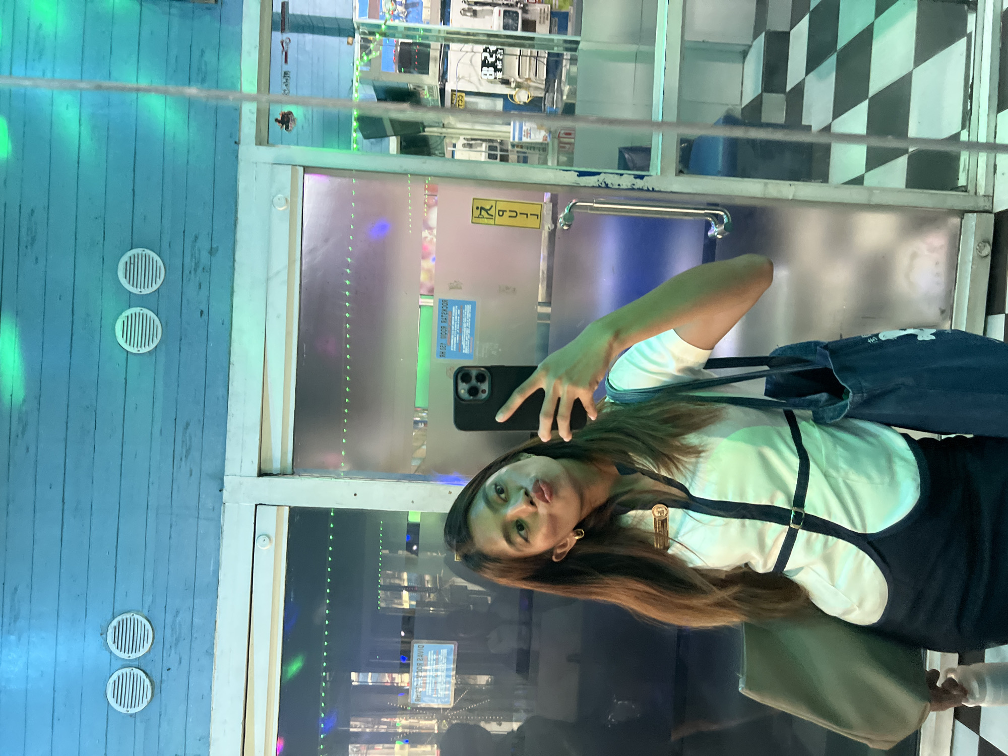 This is a vertically oriented, full-body, outdoor selfie taken by a young woman in what appears to be an entertainment or arcade setting in Davao City, Philippines. The woman is facing the camera, puckering her lips, and holding her phone to take the photo. She has long, brown hair and is wearing a white top with black straps and a black skirt. She is also carrying a light green bag over her shoulder.
The background shows a glass door with reflections, revealing a checkered black and white floor pattern behind the woman and a street scene with lights and what appear to be arcade games or vending machines. To the left of the door is a blue wooden wall with two circular white vents. Green lights are cast on the wall and reflected in the glass. A yellow sign with a running figure and the word "PUSH" is visible on the door. Another sign in blue text is also visible on the door. The overall impression is a playful and vibrant atmosphere. The time of day is unclear due to the artificial lighting and reflections, but the environment suggests it is likely evening or night.