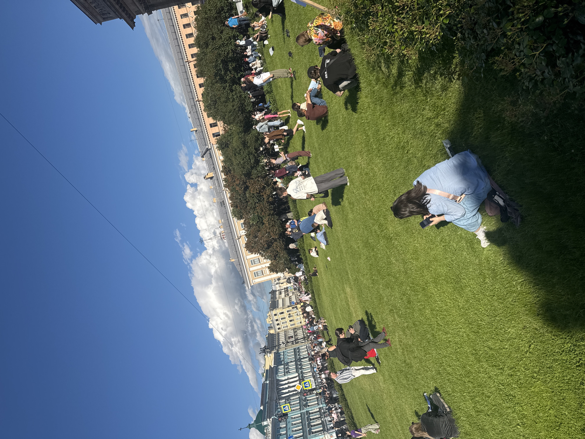 This image depicts a sunny day in Saint Petersburg, Russia, likely on the Field of Mars. A large gathering of people are spread out on a vibrant green lawn, some sitting, some standing, and some walking. The sky above is a brilliant blue with scattered white clouds. In the background, several ornate historic buildings with yellow and blue facades line a street filled with more people and trees. The overall scene suggests a public gathering or leisure activity in an urban park.