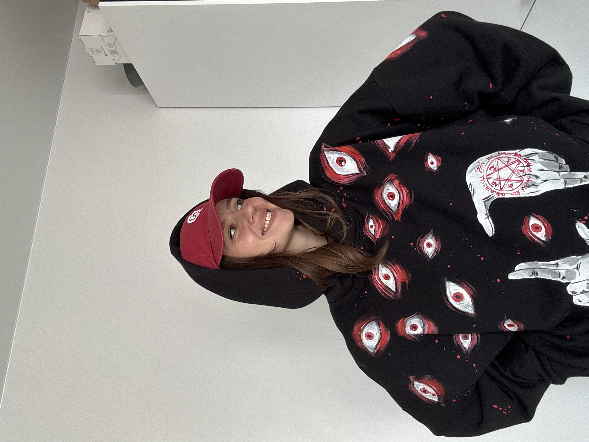 A young woman wearing a red baseball cap and a black hoodie with white eyes and hand prints smiles at the camera. The background is a white wall and a portion of a white ceiling with a mechanical fixture attached. The location is undetermined.
