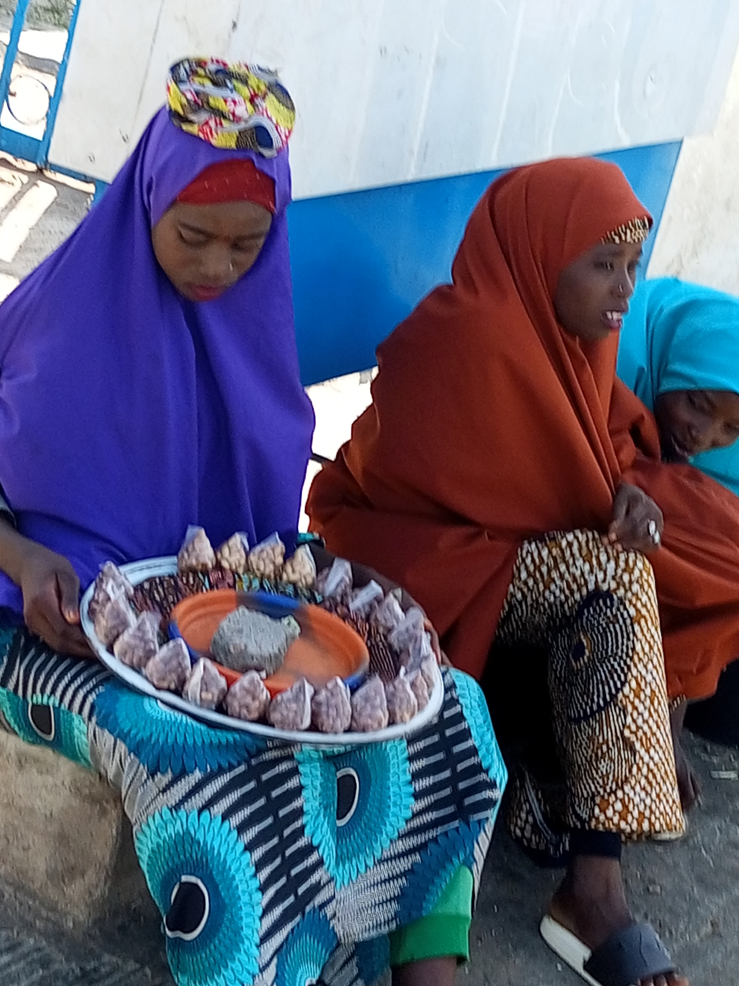 Three young females, likely vendors, are seated outdoors in Gombe, Nigeria, with a large, pale blue and white boat-like structure behind them. The female on the left wears a vibrant purple hijab and a colorful patterned headscarf, with a red underscarf. She is holding a circular tray filled with small, wrapped items and a piece of greyish material in the center. Her attire includes a blue and black patterned skirt. The central female wears an orange hijab with a patterned trim, and her legs are clad in patterned trousers featuring a geometric design in brown, white, and black. She appears to be looking down at something in her lap. The third female, to the right, is partially visible, wearing a bright teal hijab and looking downwards. The ground appears to be a mixture of sand and small stones. The overall impression is a candid street scene, likely depicting a commercial exchange or preparation for one.