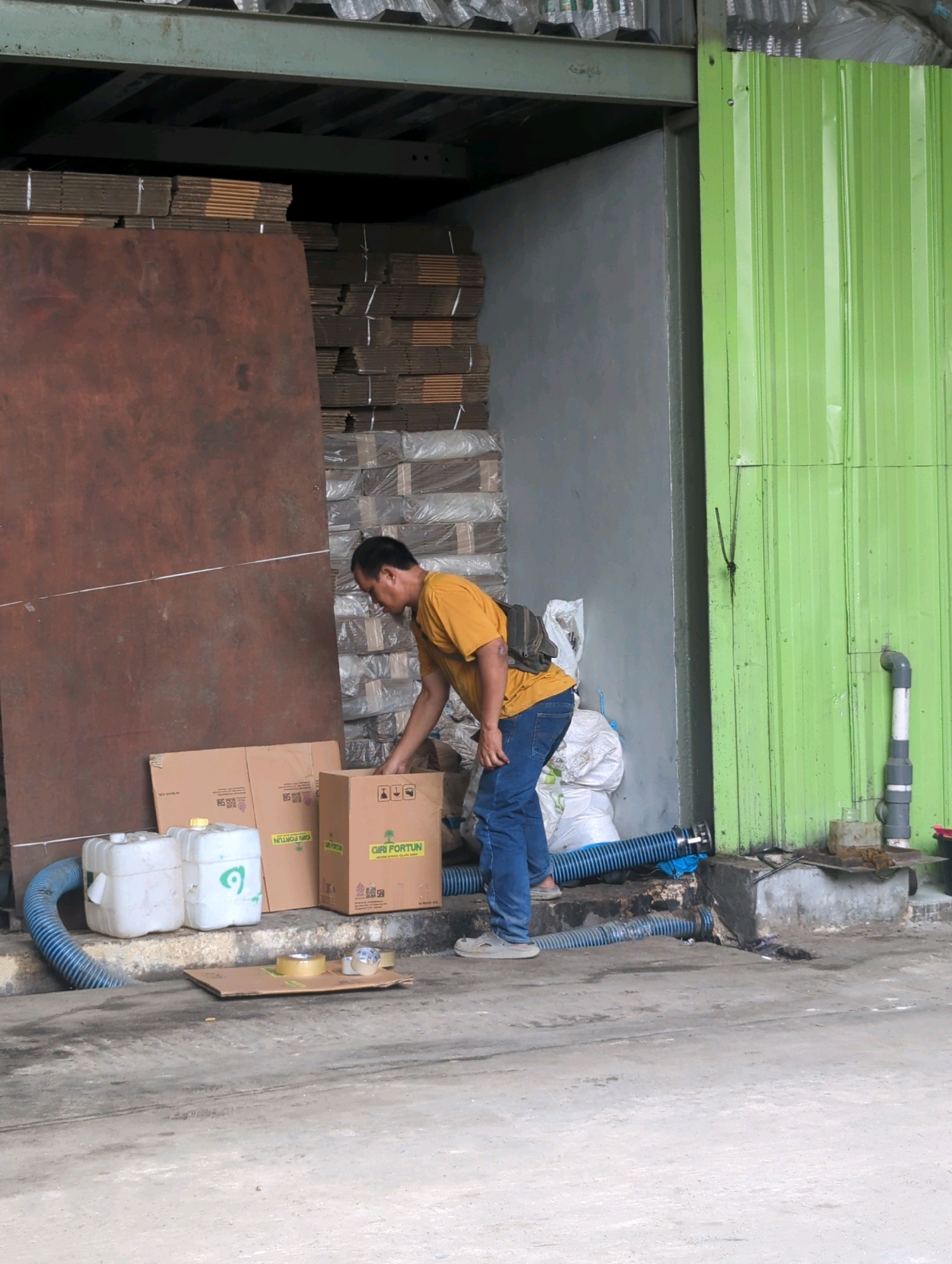 The image depicts an outdoor industrial or storage area in Batu Belaman, Indonesia. A man in a yellow t-shirt and blue jeans is bending down, reaching into a cardboard box. He has a backpack slung over his shoulder. In front of him are two white plastic containers with yellow caps, and two brown cardboard boxes, one with the label "CIRI FORTUN". A blue corrugated pipe runs along the ground, connecting to one of the containers and appearing to lead towards a drain. Stacked in the background are numerous flat, brown cardboard sheets, wrapped in plastic. To the right, a corrugated green metal wall stands behind a concrete structure with plumbing. The ground is a rough, concrete-like surface.
