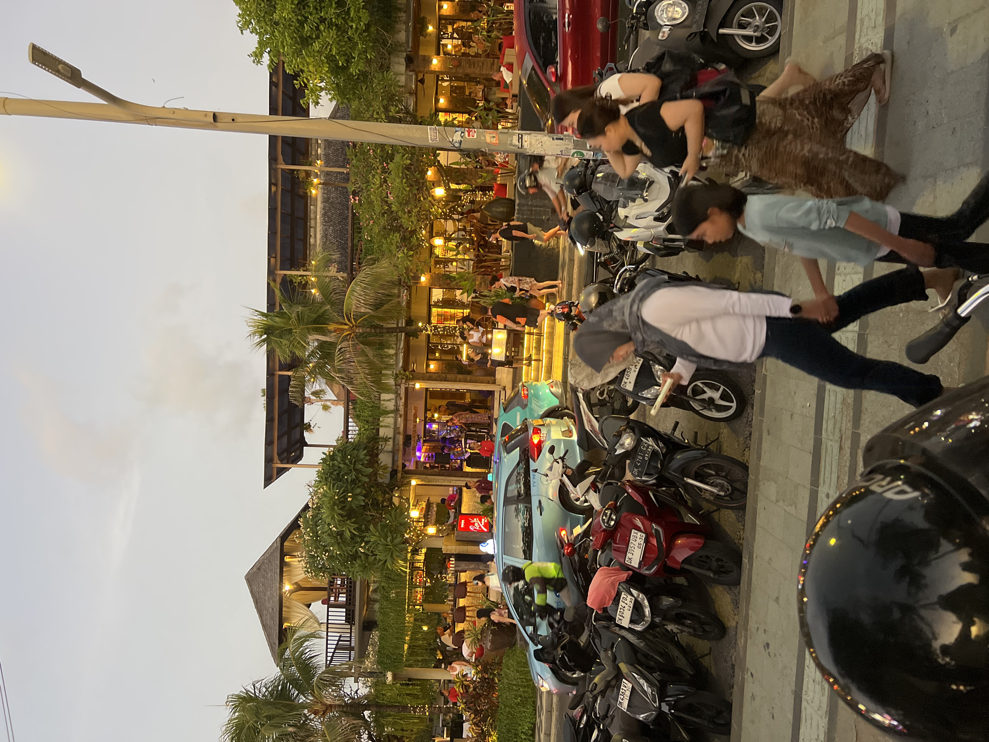 The image captures a street scene in Kuta, Indonesia, likely during the evening. A row of parked motorcycles is visible in the foreground, with several people walking along the sidewalk. In the background, a restaurant or bar area is illuminated with warm lighting, with patrons seated at tables and standing near a bar. Palm trees and tropical foliage add to the ambiance. A light blue car, possibly a taxi, is driving on the street. The sky is overcast with hints of smoke or haze. The overall scene depicts a bustling commercial and social hub in a tropical city.