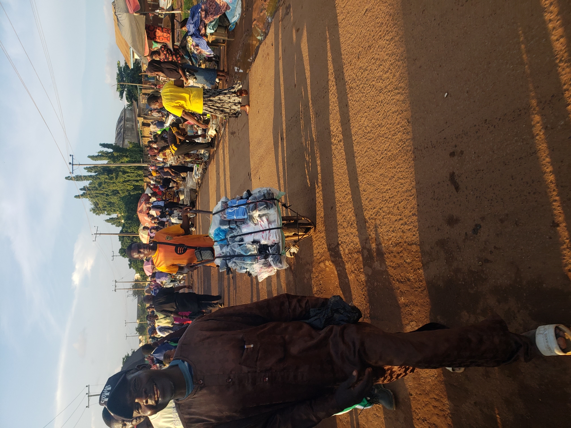 The image depicts a bustling market scene in Kakuri Gwari, Nigeria. In the foreground, a man wearing a brown tunic and a black cap smiles at the camera, holding a black plastic bag in his left hand. He is walking towards the viewer on a dirt road. Behind him, a man in an orange shirt pushes a wheelbarrow laden with neatly packed plastic bags. Numerous people are visible in the background, engaged in various market activities, with stalls and umbrellas suggesting commerce. The environment is an outdoor market with a dusty, unpaved road and a clear sky with scattered clouds. The overall impression is of a lively community gathering.