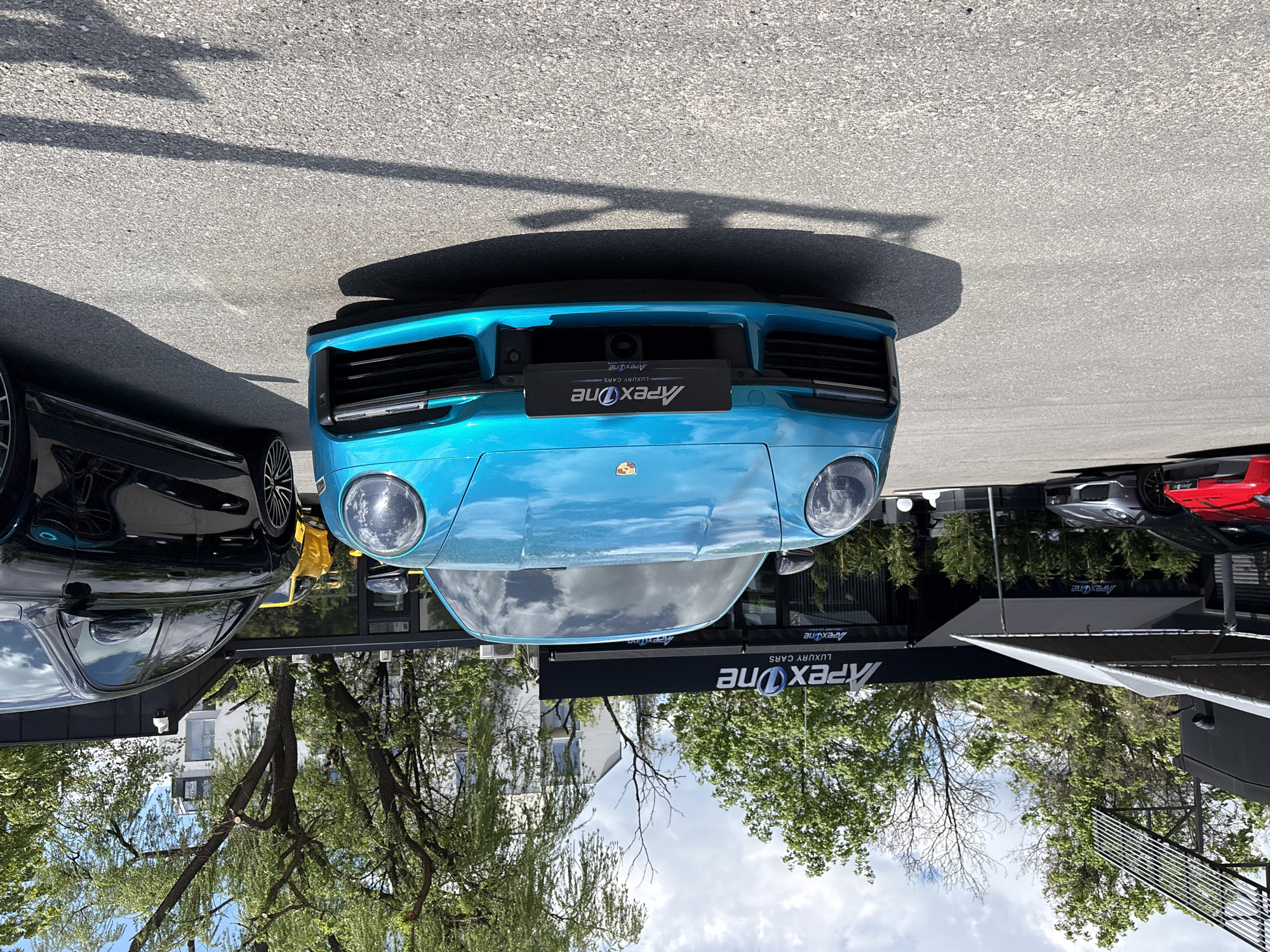 The image shows the front view of a turquoise Porsche 911 with an "Apex One Luxury Cars" license plate frame. The car is parked outside the Apex One dealership. There are other luxury cars parked around the turquoise car, including a black car on the left. Trees are visible in the background, and the sky is cloudy. The city is Sofia, Bulgaria, based on the Apex One dealership location.