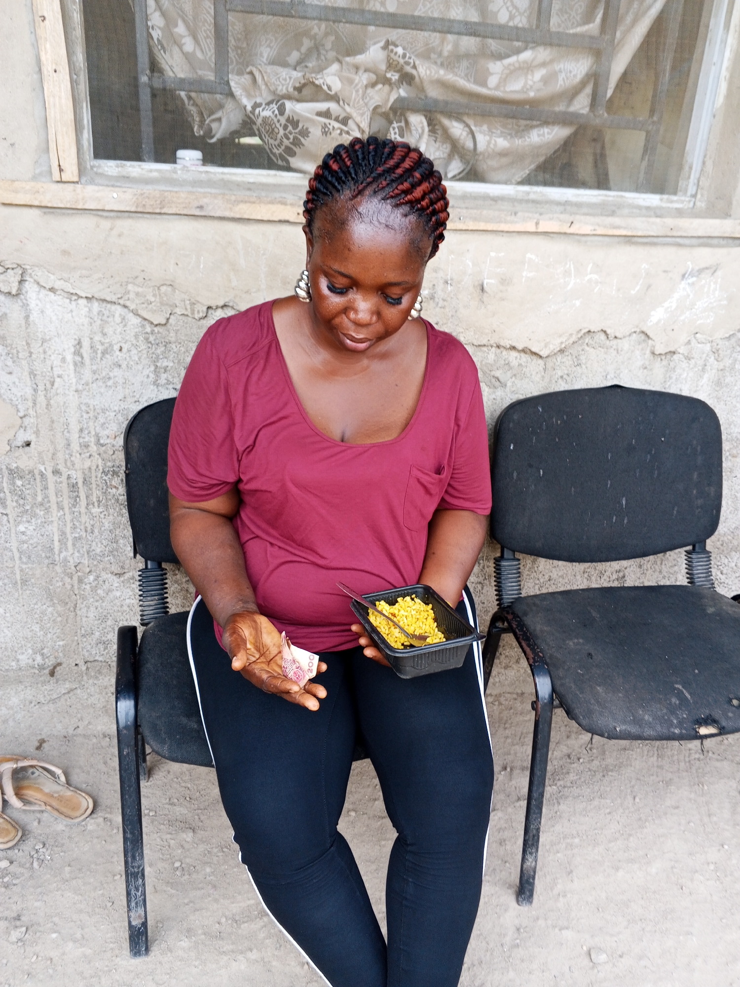 In Adonkolo, Nigeria, a woman is sitting on one of two black chairs. She is holding a black container of corn and a 200 Nigerian Naira note. She is wearing a maroon t-shirt and black pants with a white stripe. Sandals are on the ground near the chair she is sitting on. Behind her is a dilapidated wall with a window.