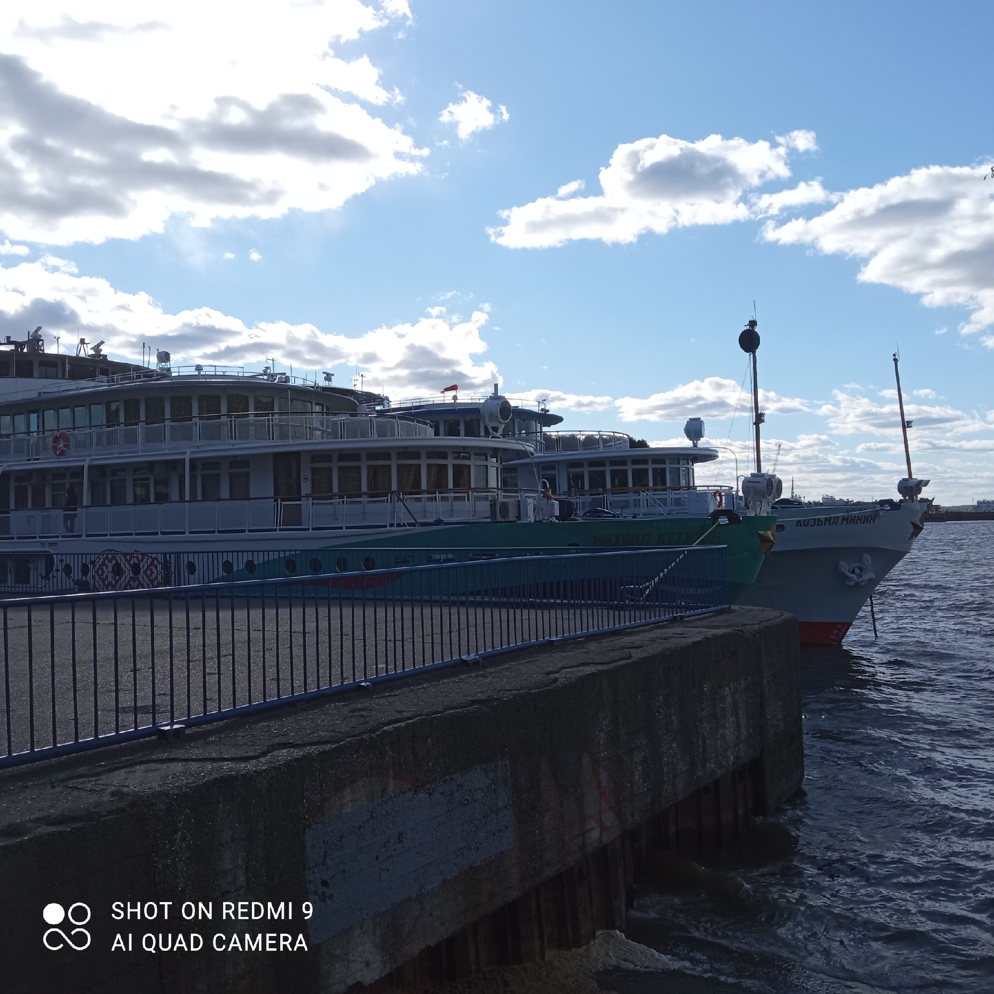 The image depicts a waterfront scene in the city of Chaikovsky, Russia, featuring several river cruise ships docked along a concrete pier. The ships, varying in size and design, are lined up parallel to the pier. The ship closest to the viewer has the words "КОЗЬМА МИНИН" printed on the hull. The ships are multi-decked, with multiple windows and balconies. A dark metal railing runs along the edge of the pier. The water is choppy, with visible wave patterns. The sky is partly cloudy, with patches of blue visible between the clouds. The lighting suggests a sunny day. The words "SHOT ON REDMI 9 AI QUAD CAMERA" are visible in the lower left corner of the image.