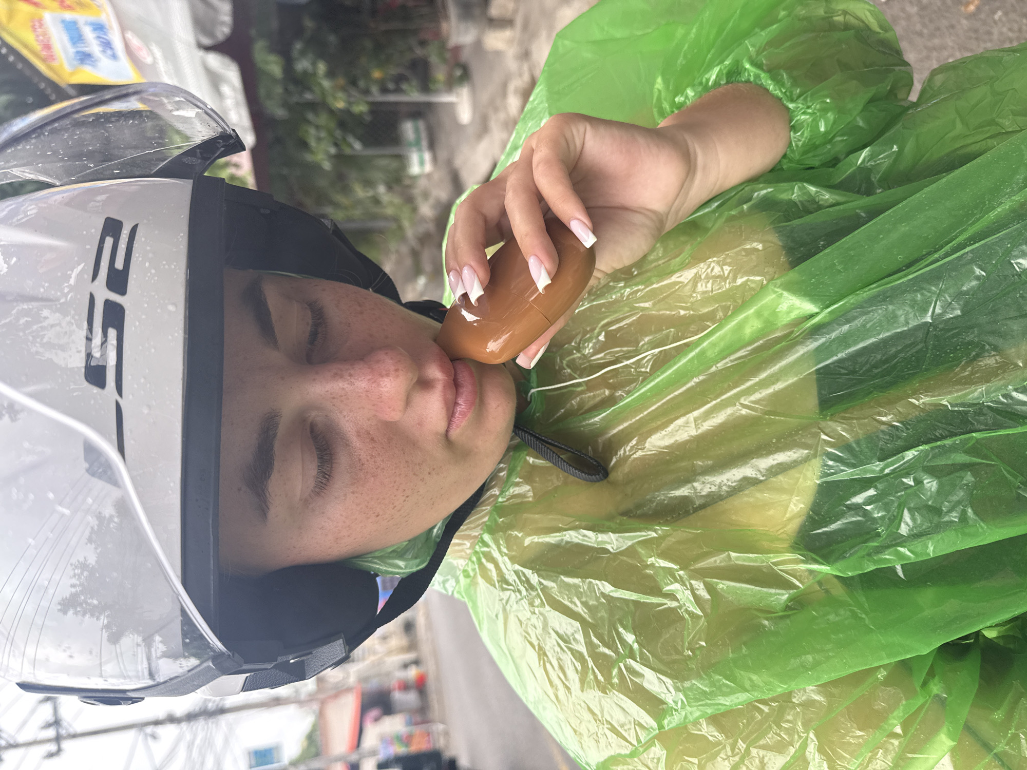 The image shows a close-up, from the chest up, of a young Caucasian woman wearing a white LS2 helmet with a clear visor, and a bright green plastic raincoat. The woman has a light complexion with freckles across her nose and cheeks, and her eyes are closed. She is holding a small, oval-shaped brown object to her lips with her right hand. Her fingernails are long and painted white with a French manicure. The background is blurred, but appears to be an outdoor urban setting with buildings, power lines, and foliage, consistent with the city of Nha Trang, Vietnam. The atmosphere suggests a cloudy or rainy day, as indicated by the water droplets on the helmet visor and the plastic material of the raincoat. The scene appears to be a spontaneous moment captured while the woman is on the go, possibly riding a motorbike.